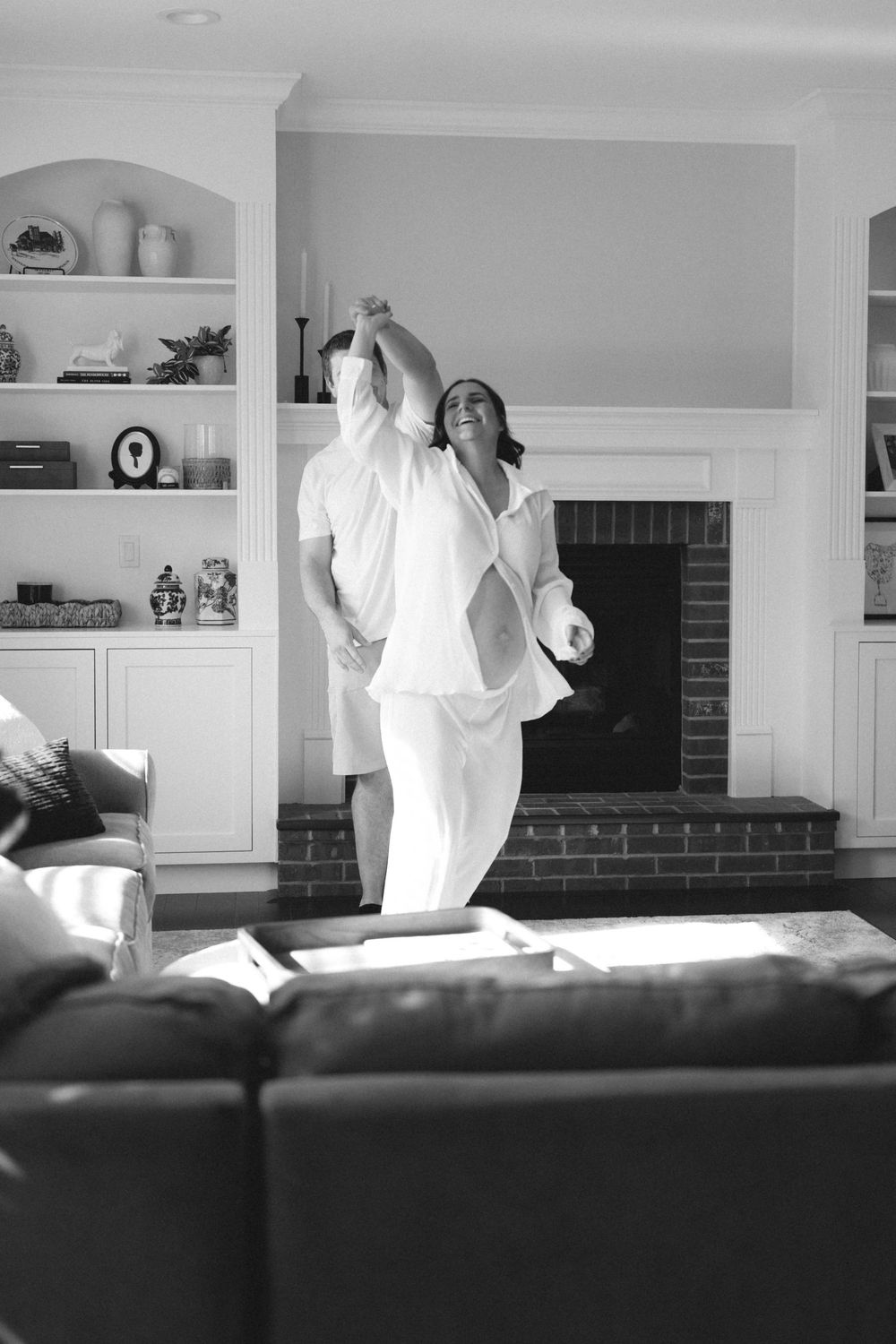 Black and white photo of a pregnant woman dancing joyfully in an elegant living room with fireplace.