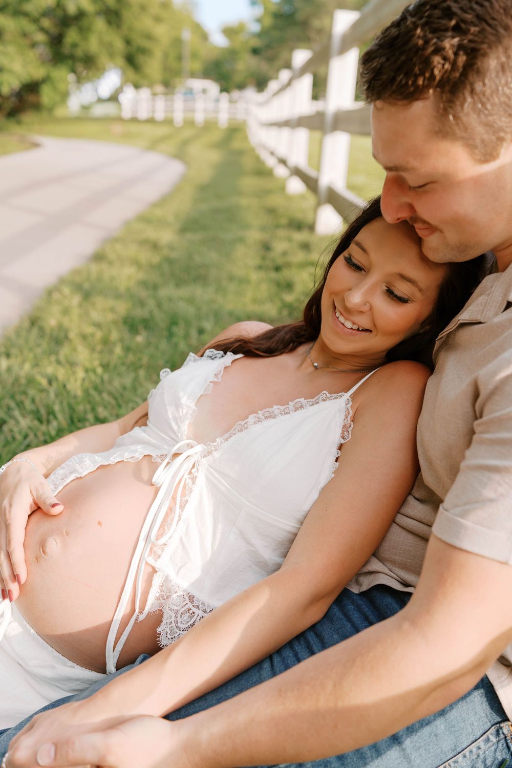 Couple sitting by white fence, man nuzzling pregnant woman as she smiles at bump.