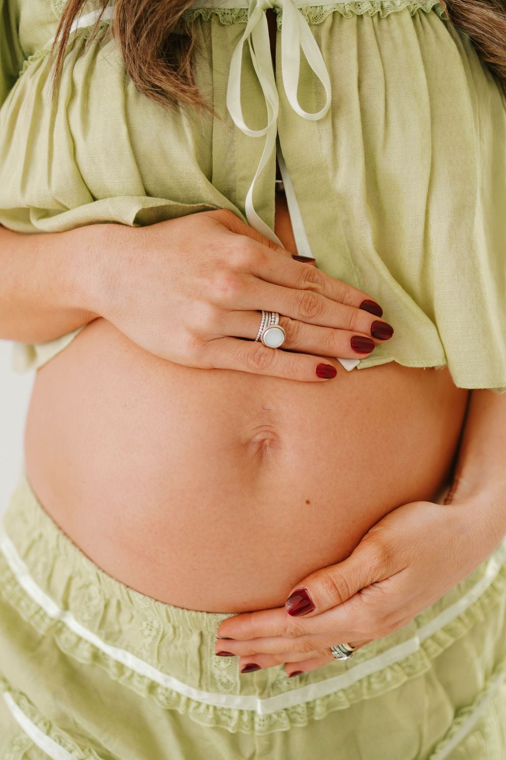 Pregnant woman in green crop top cradling baby bump with manicured hands and diamond ring.