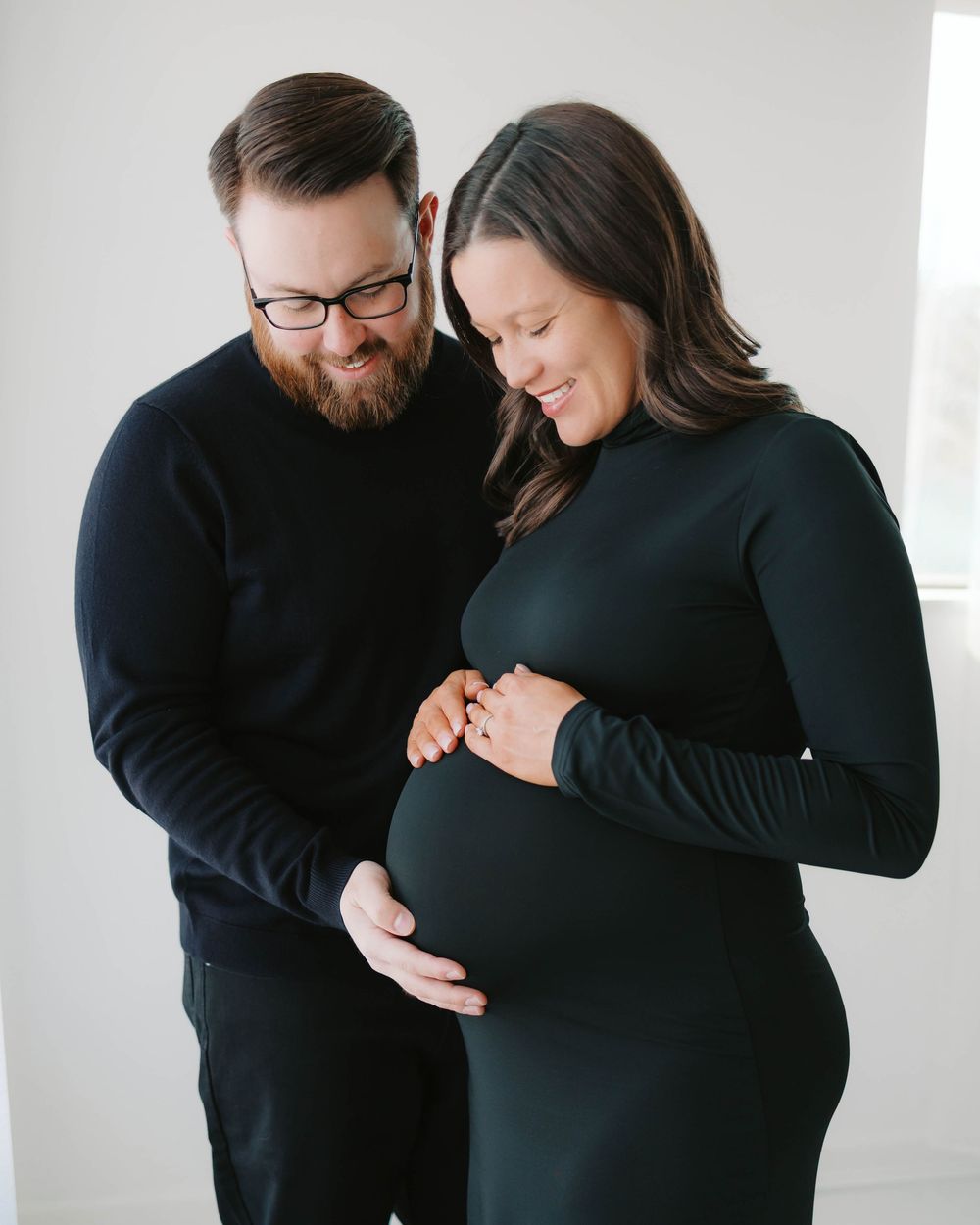 Couple in matching black outfits tenderly holding pregnant belly in bright studio.