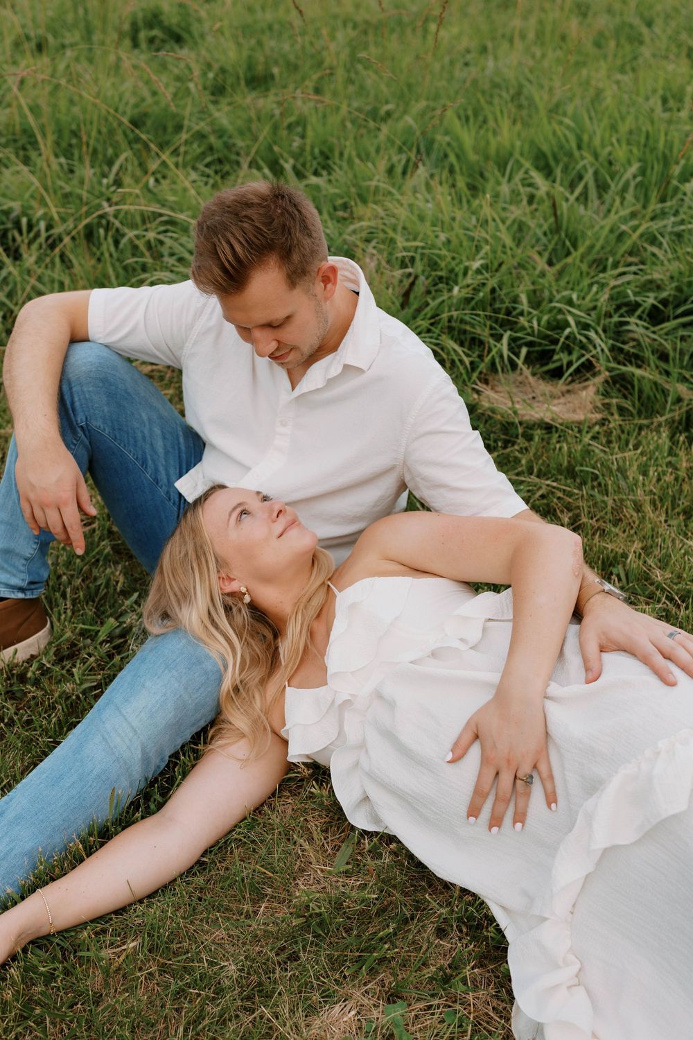 Couple relaxing in green field, woman in white dress lying across mans lap smiling.