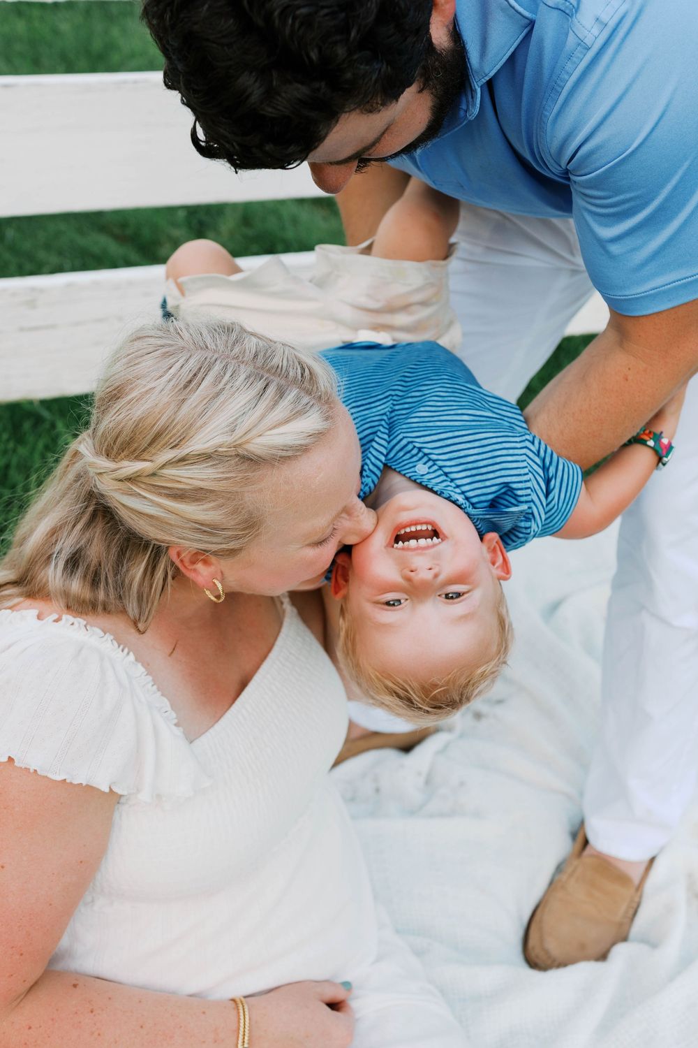 A joyful family moment with parents kissing their laughing toddler on a white bench outdoors.