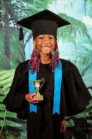 An picture of  preschool girl with pink braid, smiling at the camera for their Grade R graduation portrait, holding their graduation trophy.