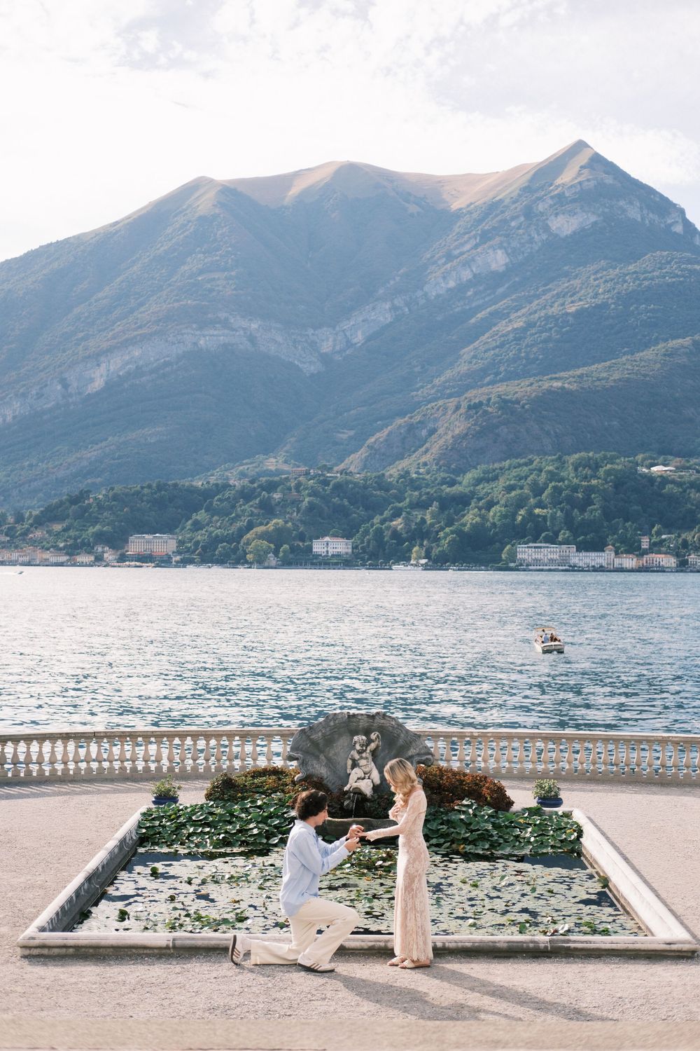 Man proposes to woman on one knee by Lake Como with stunning mountain backdrop and ornate stone balustrade.