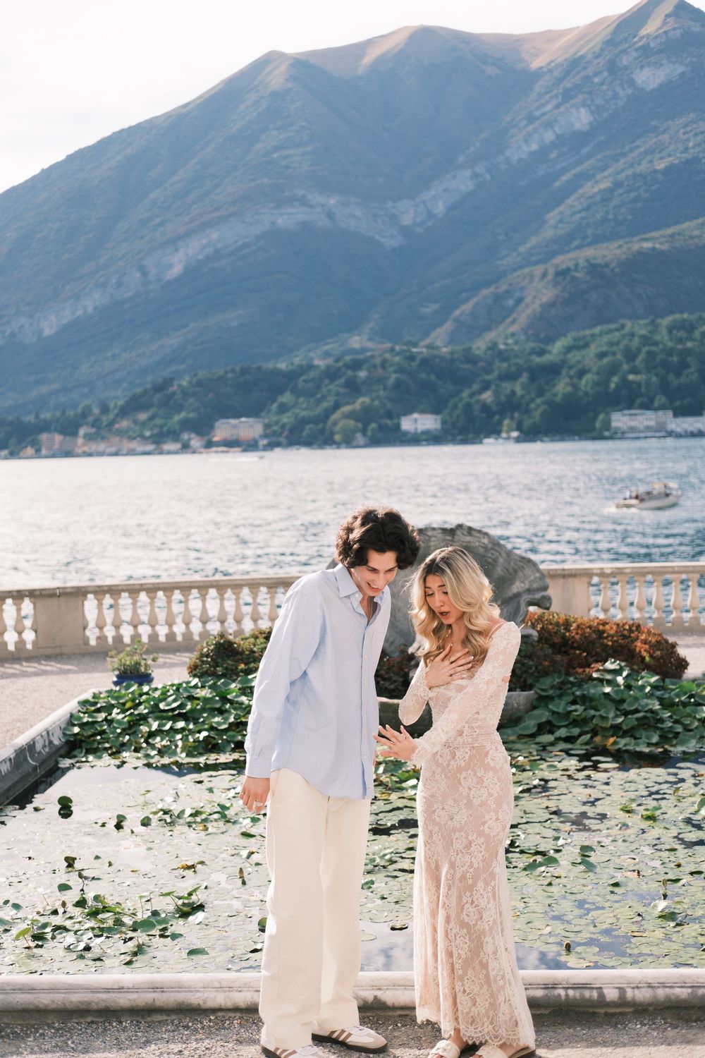 Couple in elegant attire standing on a lakeside terrace with mountain backdrop and serene water views.