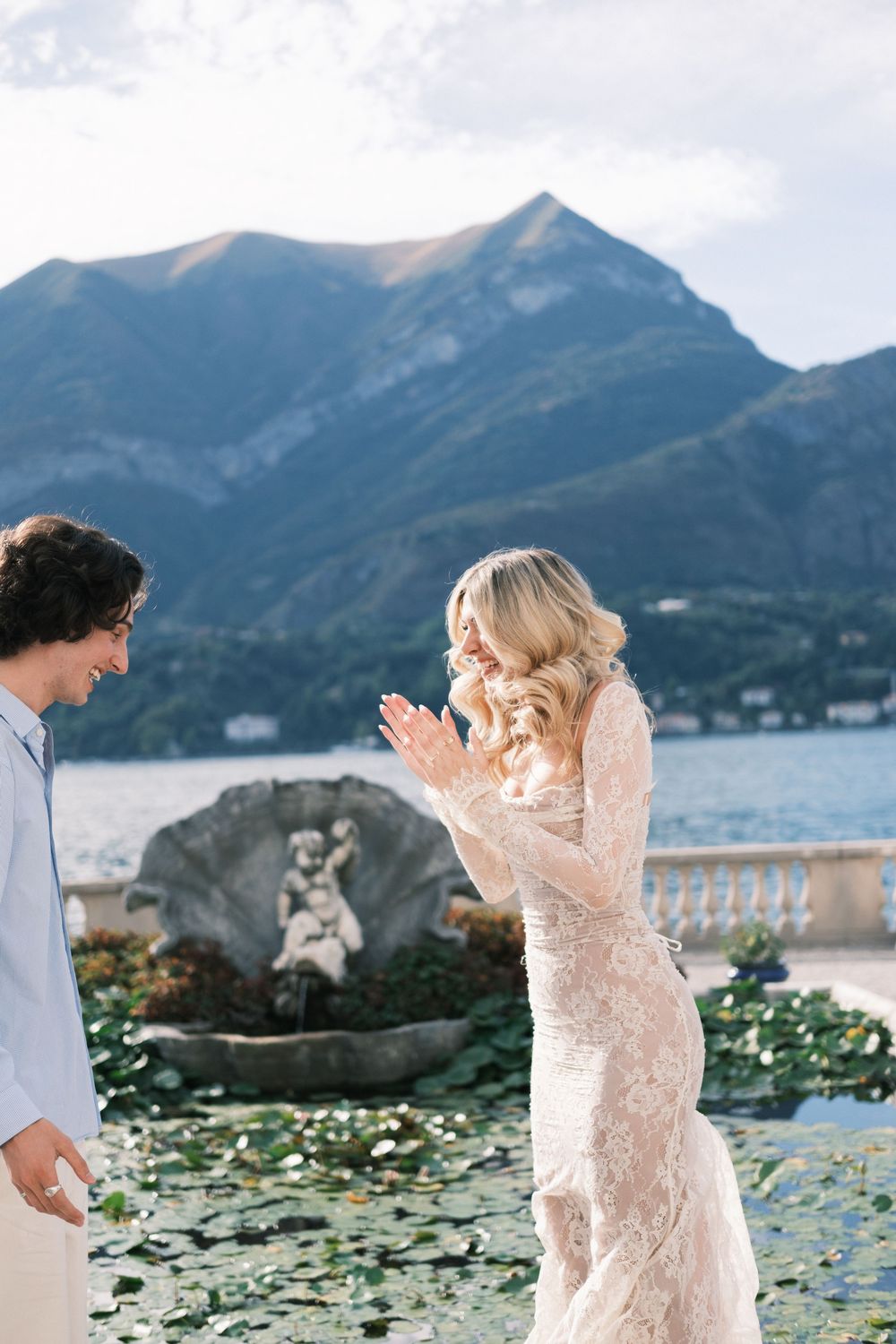 Bride in lace gown clapping joyfully with groom by Lake Como fountain, mountains backdrop.