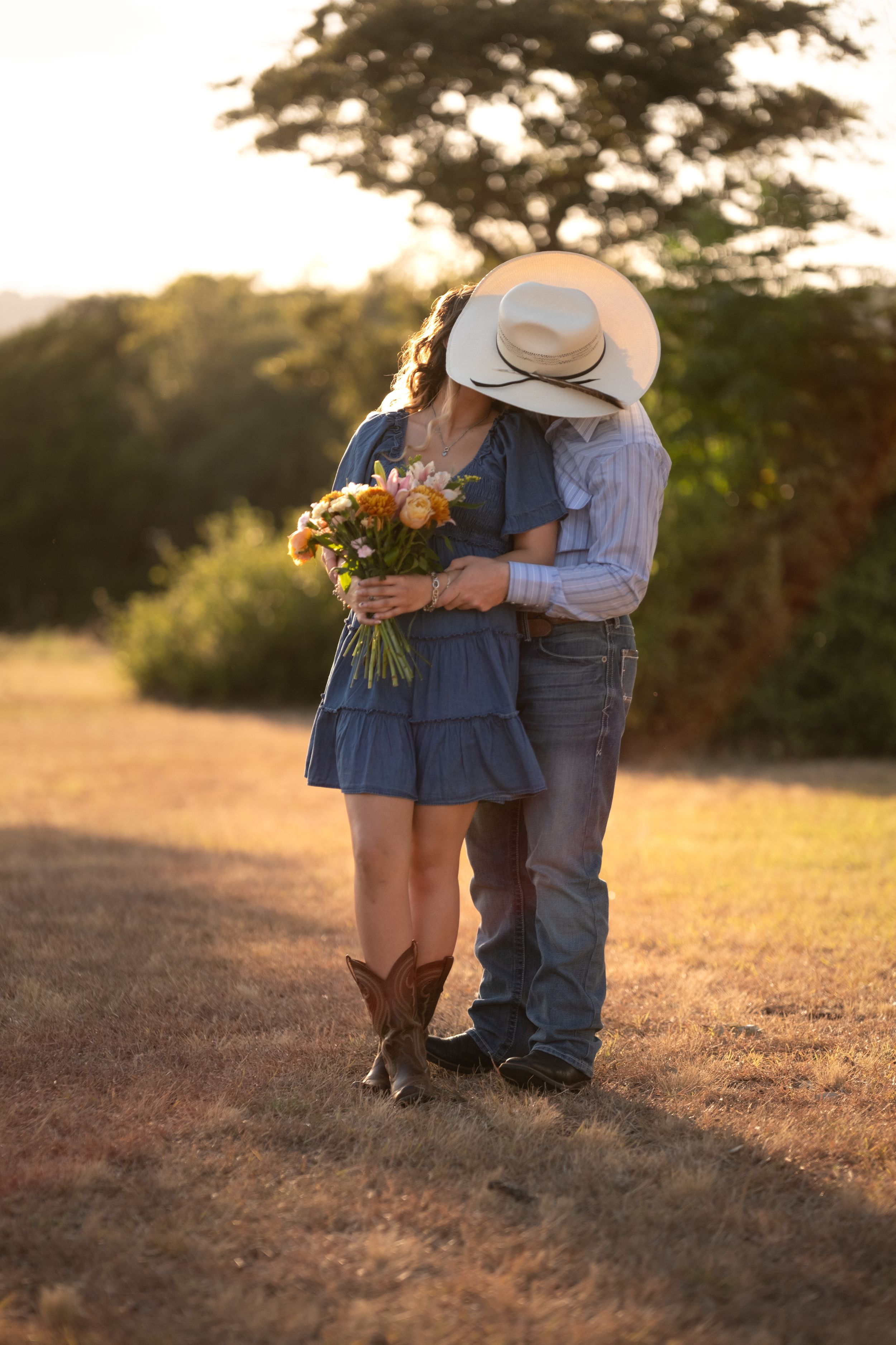 Couple kissing behind a colorful bouquet at golden hour, woman in blue dress and cowboy boots in Belton Texas