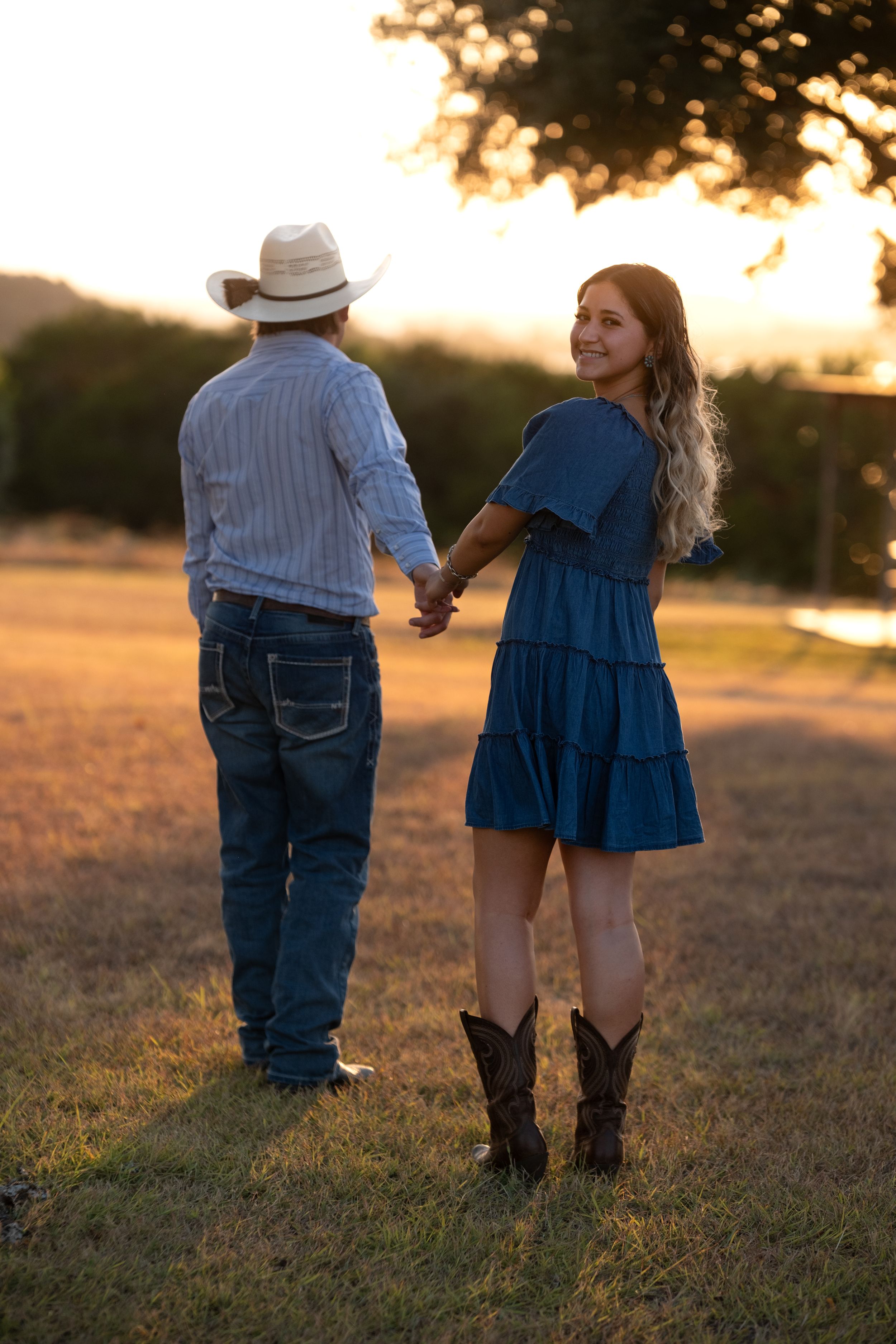 Couple holding hands at sunset, woman in blue dress and boots smiling back at camera outdoors  in Belton Texas