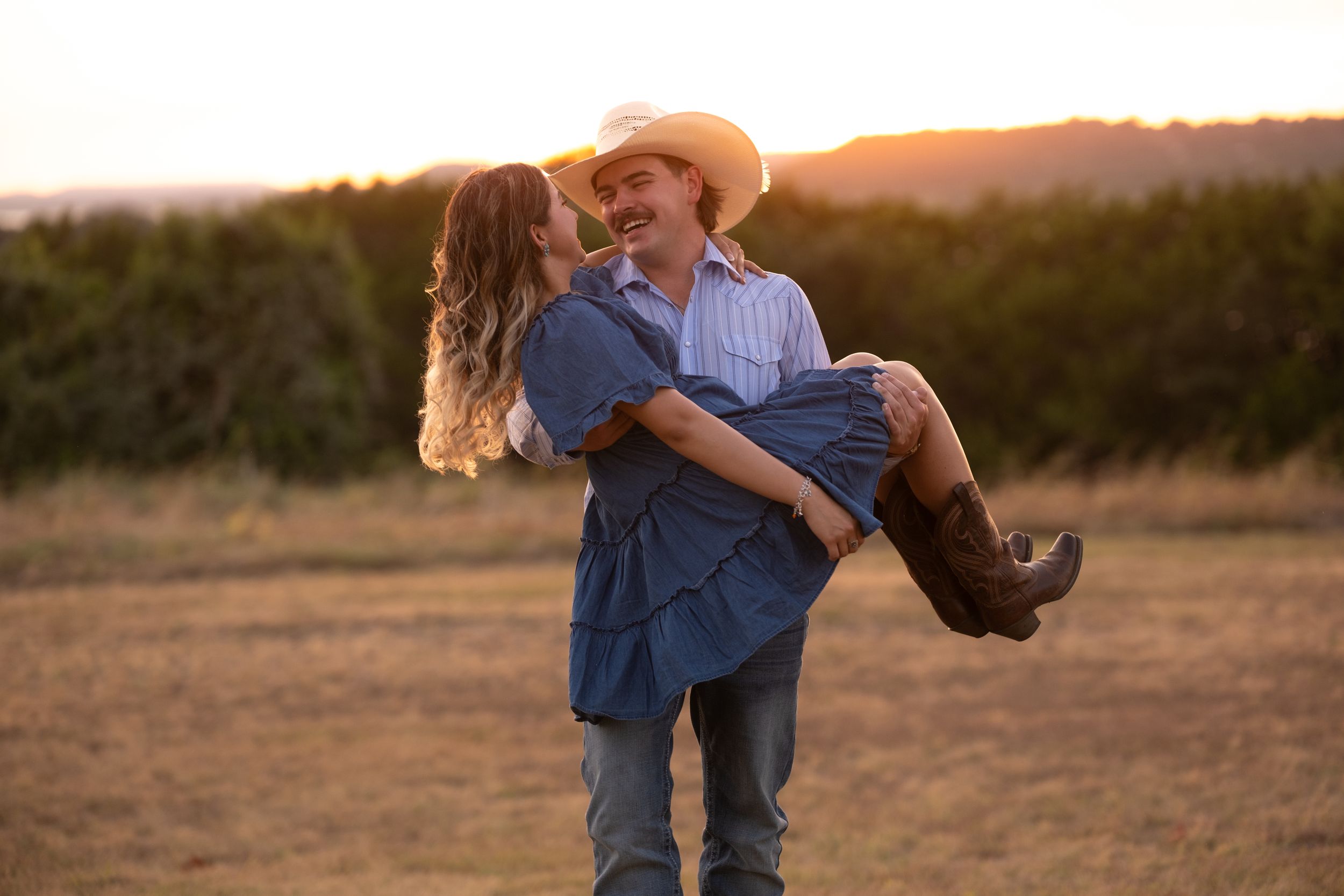 Man carrying woman in blue dress and cowboy boots at golden sunset in an open field  in Belton Texas