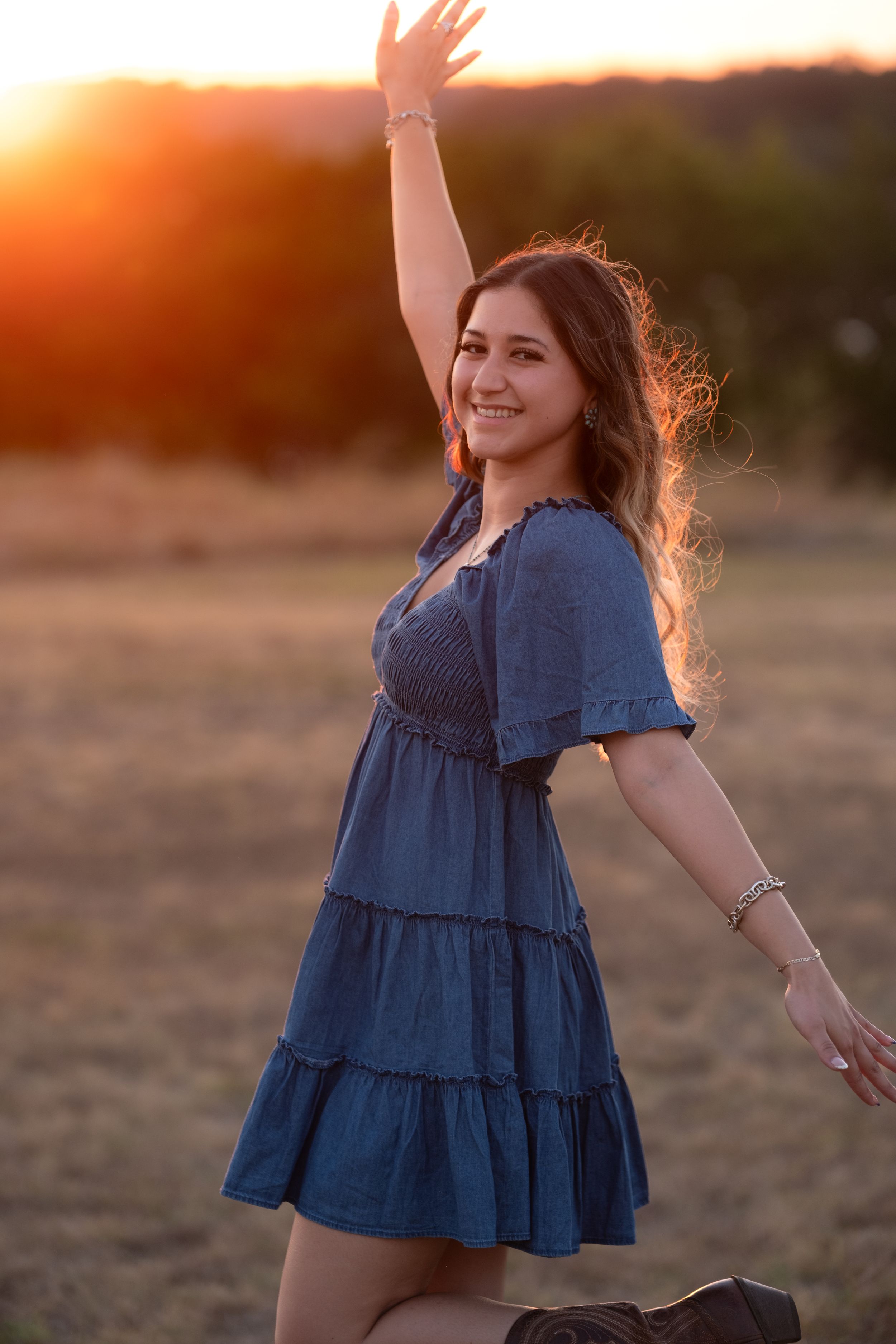 Young woman in blue tiered dress dancing joyfully at sunset in an open golden field in Belton Texas