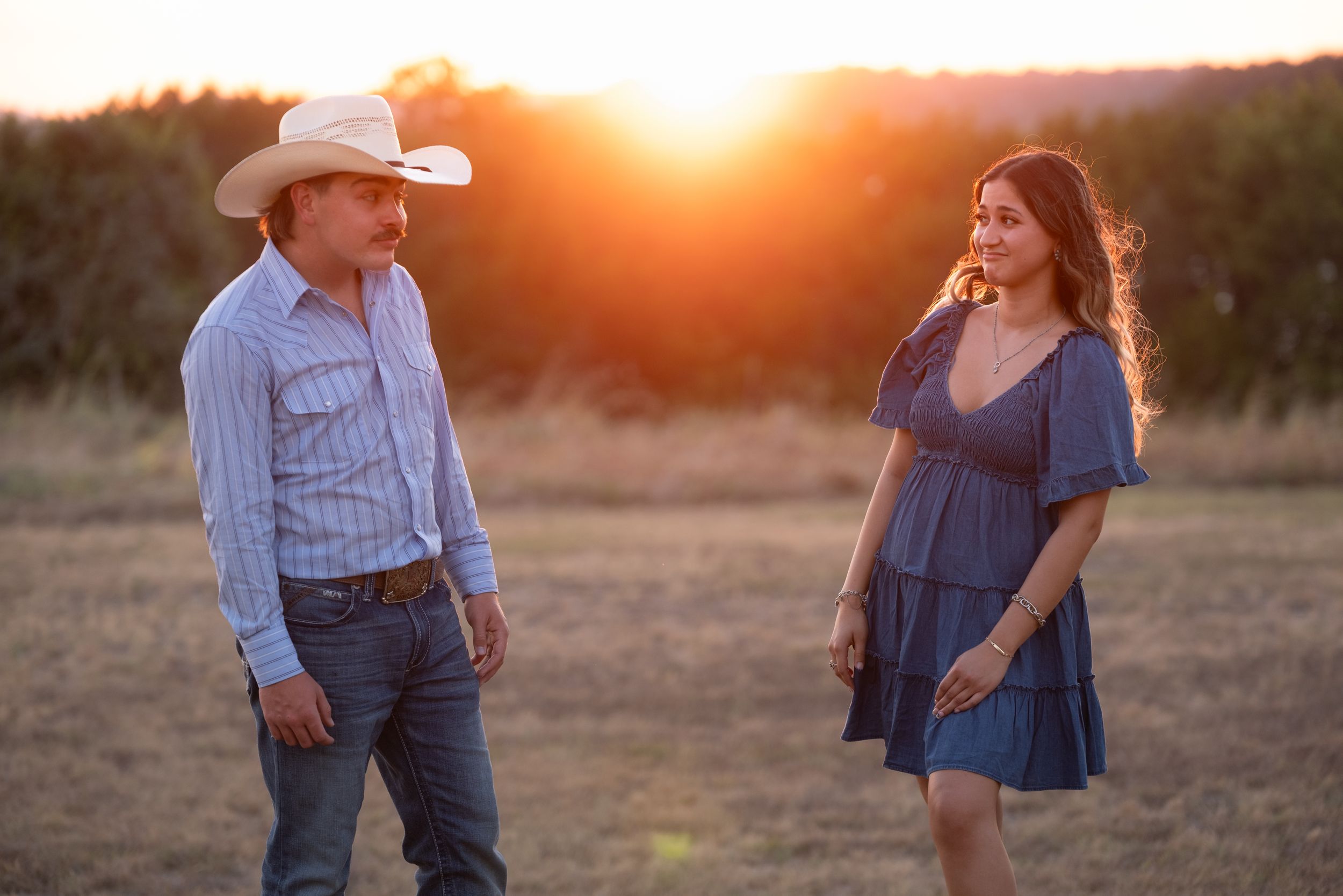 Couple standing apart in a sunlit field at sunset, man in cowboy hat and woman in blue dress  in Belton Texas