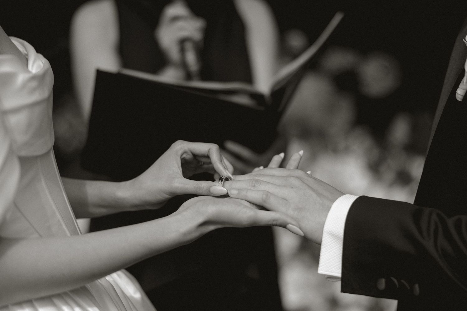 Black and white photo of bride placing wedding ring on groom's finger during ceremony.