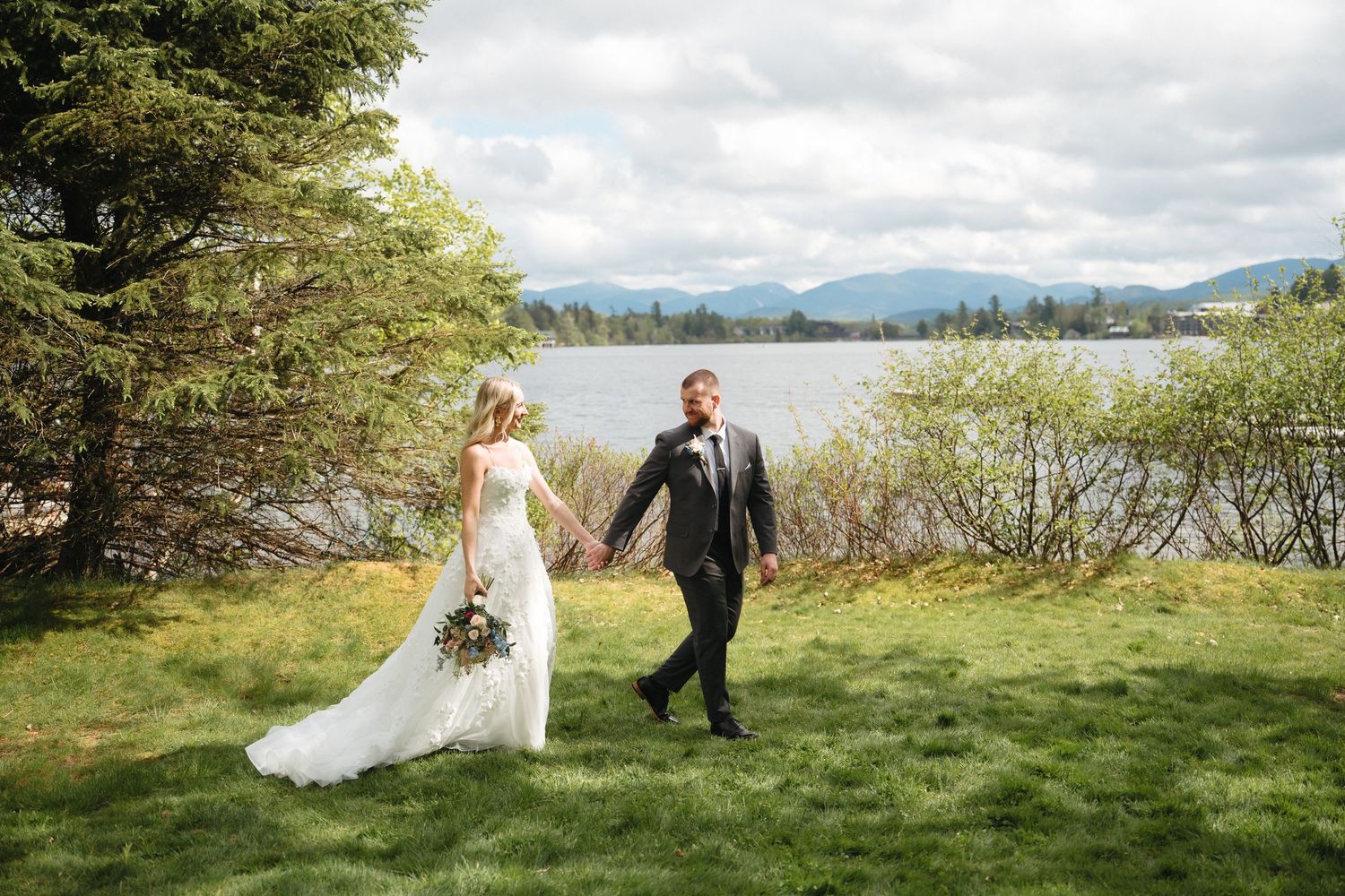 Bride and groom hold hands walking on green lawn with mountain lake backdrop on their wedding day.