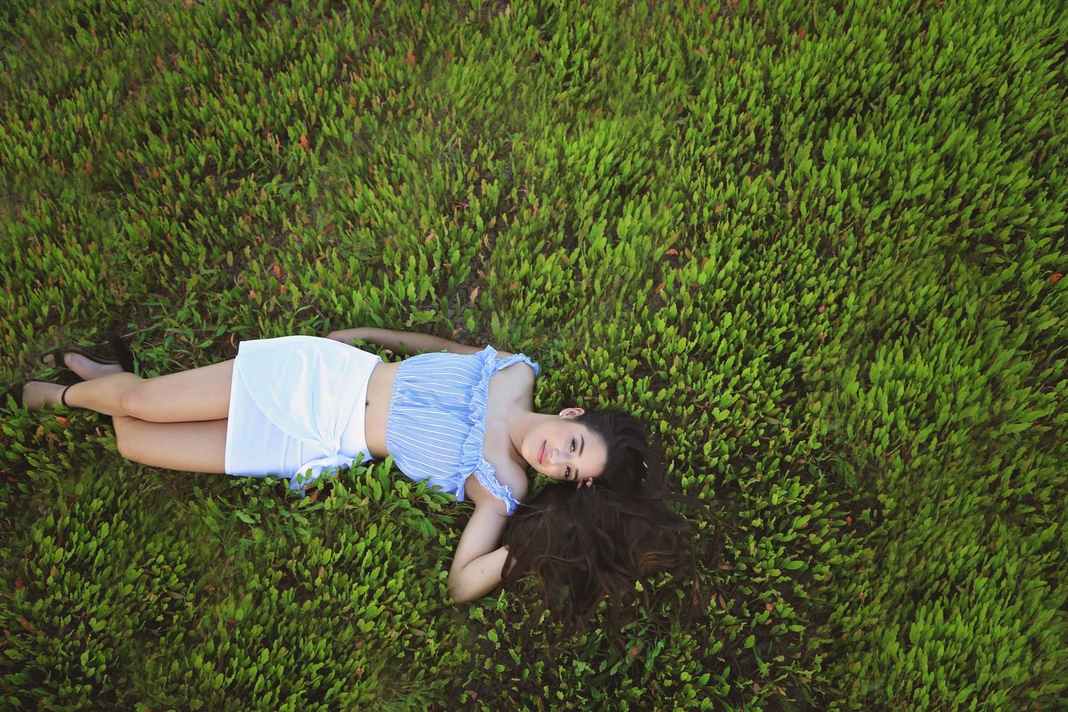 Young woman in a blue off-shoulder top and white skirt lies on a lush green grassy field, photographed from above.