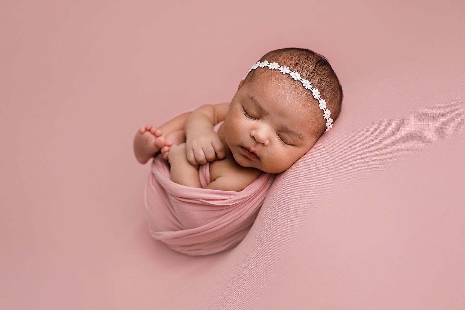 Sleeping newborn baby girl wrapped in pink swaddle with floral headband on pink background. Utah County newborn photographer