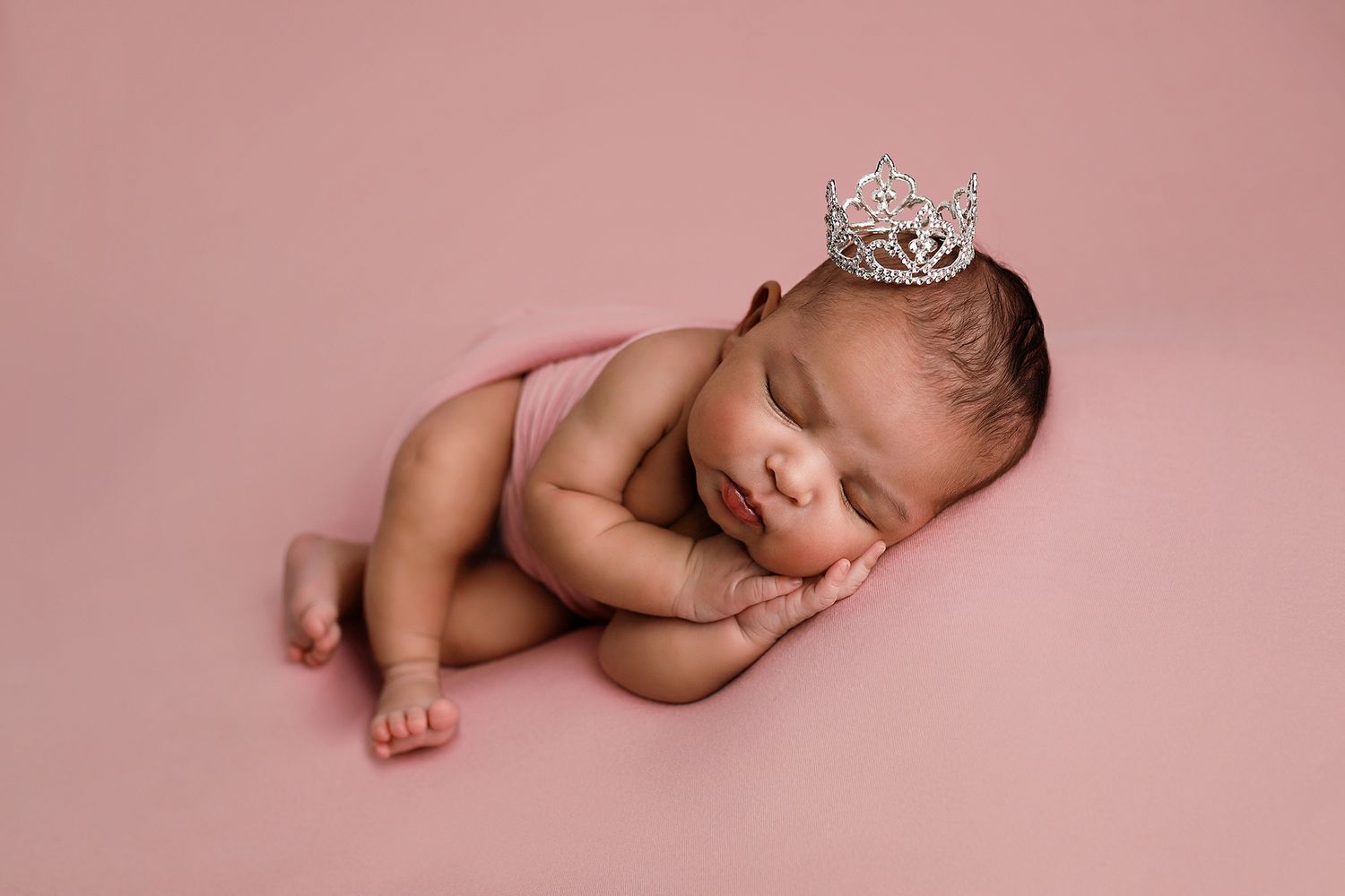 Newborn baby sleeping peacefully on pink backdrop wearing a tiny silver crown in a professional photography portrait.