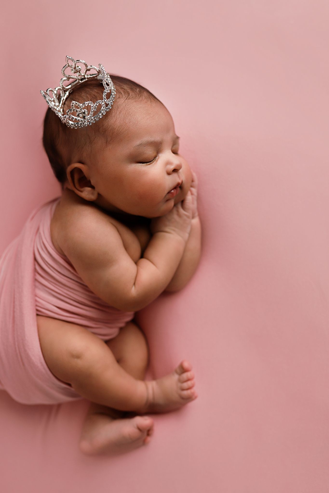 Sleeping newborn baby girl wrapped in pink fabric wearing a sparkly crown on a soft pink background. Utah County newborn photographer