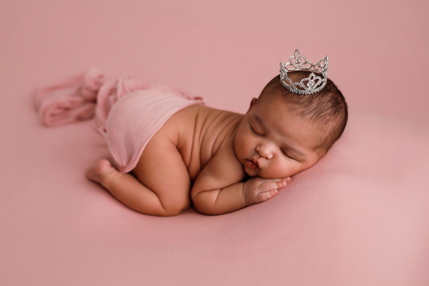 Sleeping newborn baby girl wearing a tiny tiara crown, posed on a pink blanket in a professional photo.