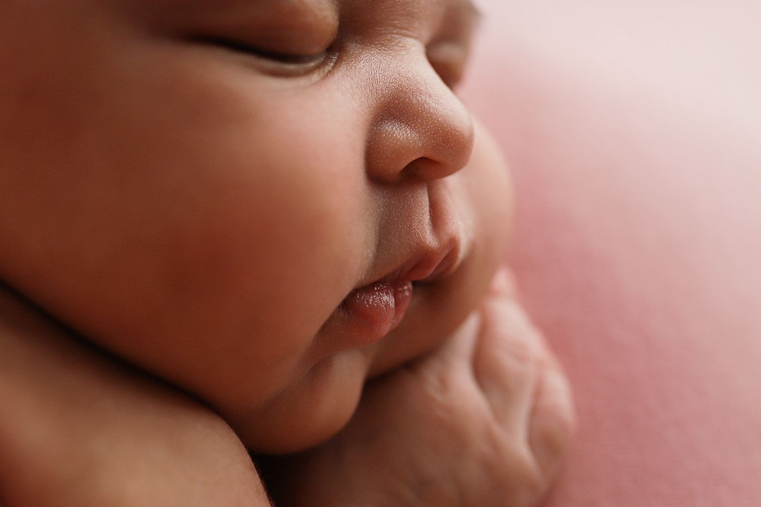 Close-up of a sleeping newborn baby with tiny hands tucked under chin on a soft pink background.