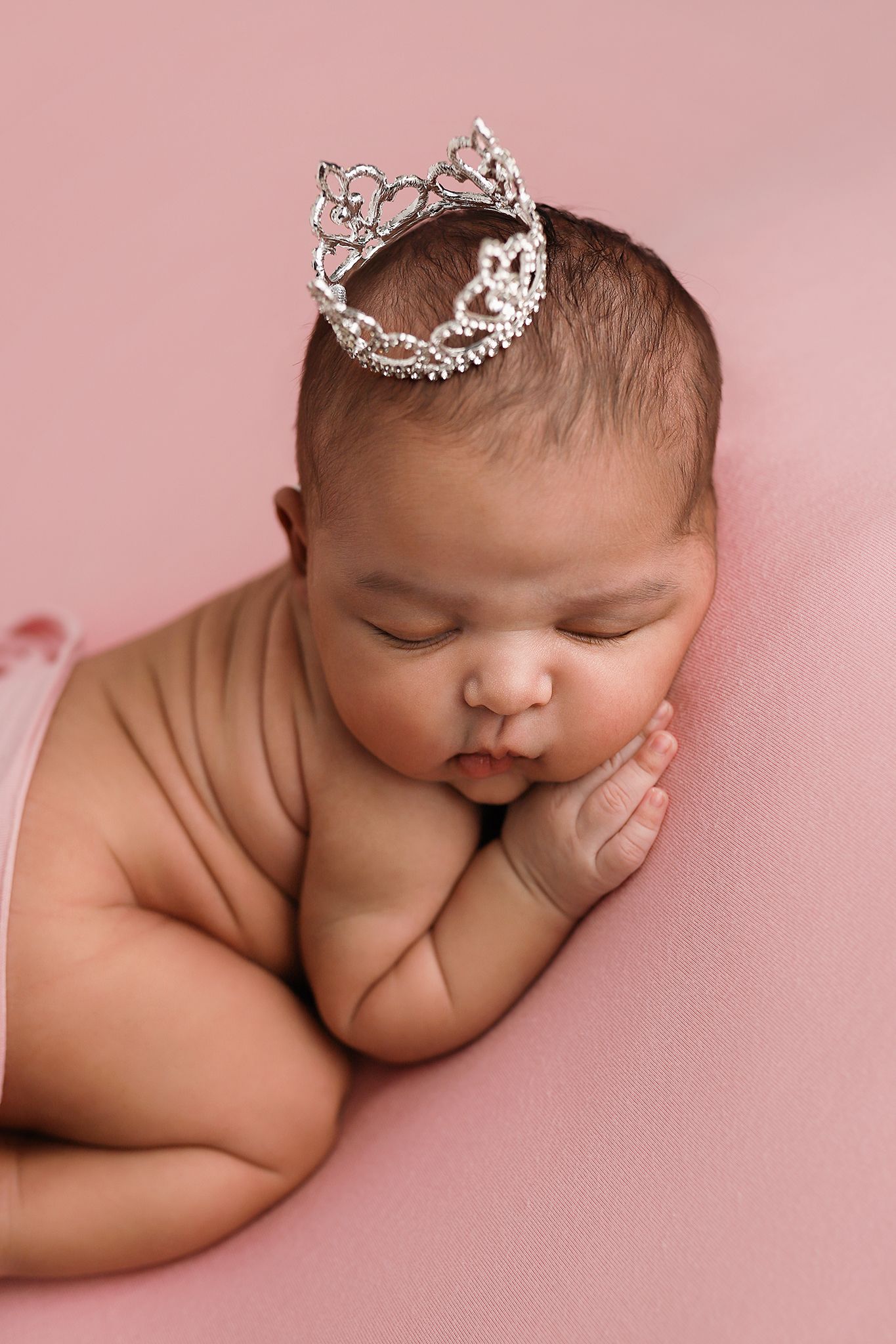 Sleeping newborn baby girl wearing a sparkling silver tiara crown on a soft pink background.
