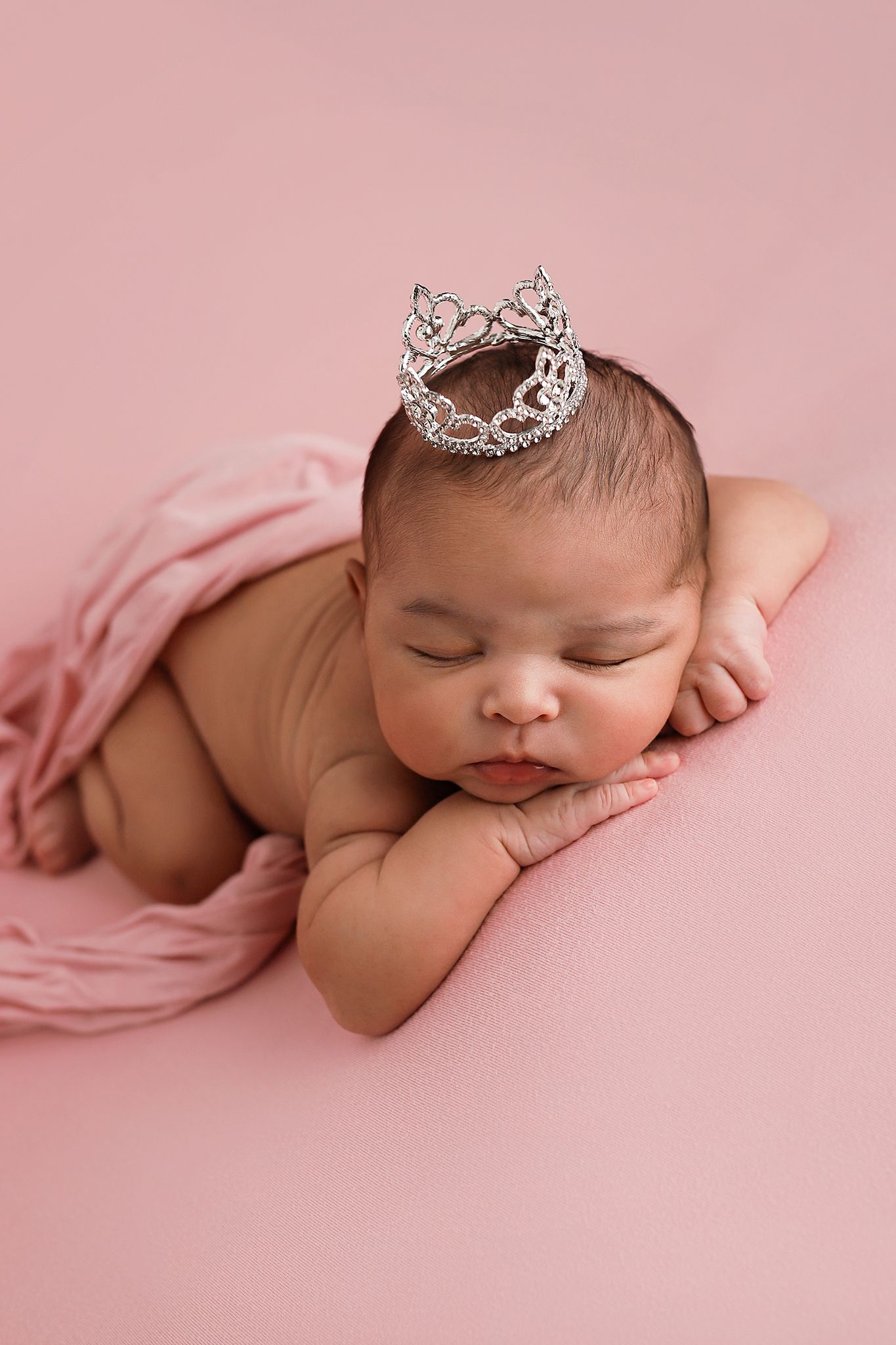 Sleeping newborn baby wearing a tiny silver tiara crown on a pink background. Utah County newborn photographer