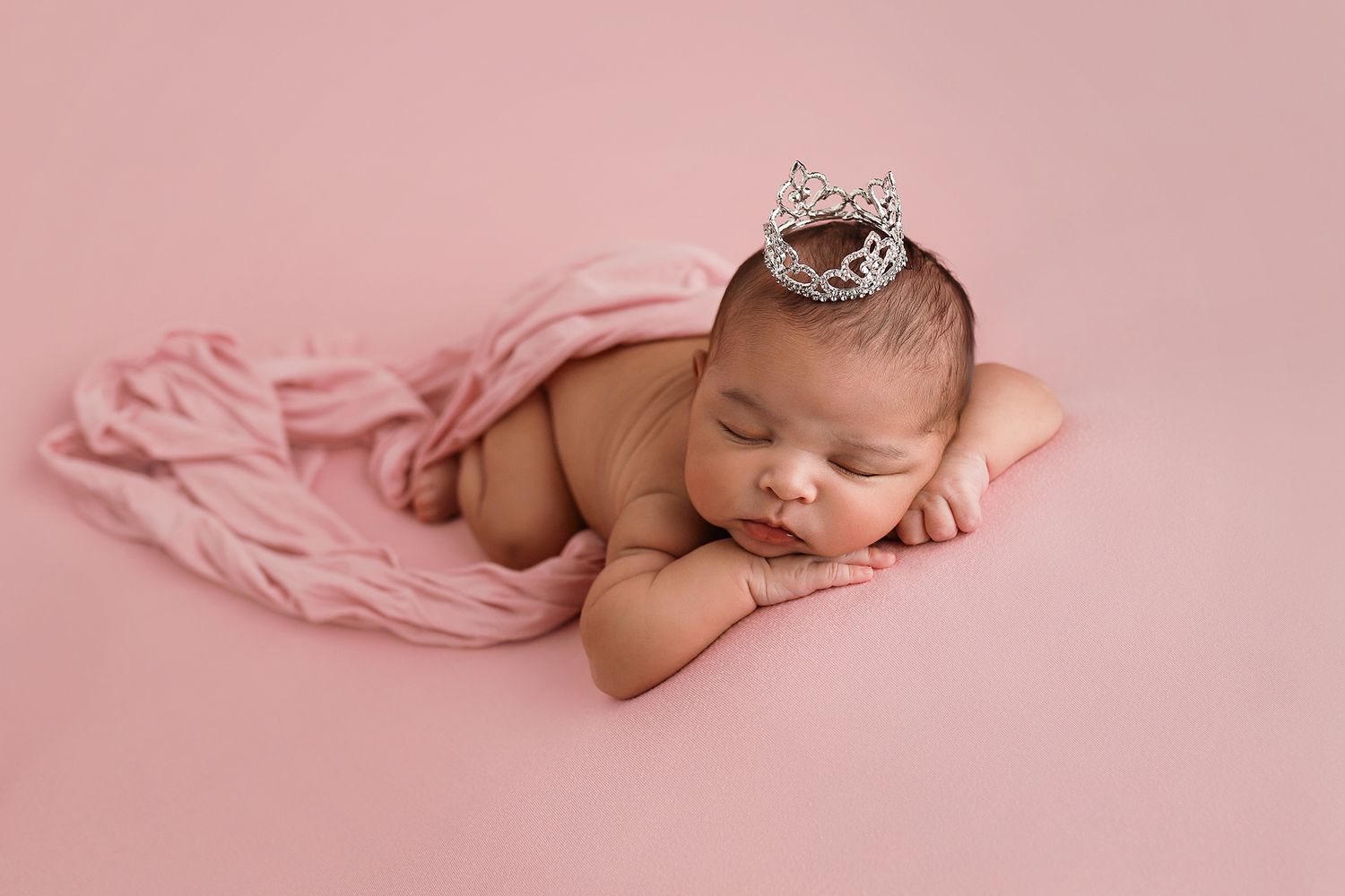 Sleeping newborn baby wearing a tiny silver crown on a pink backdrop, wrapped in a soft pink blanket. Utah newborn photography