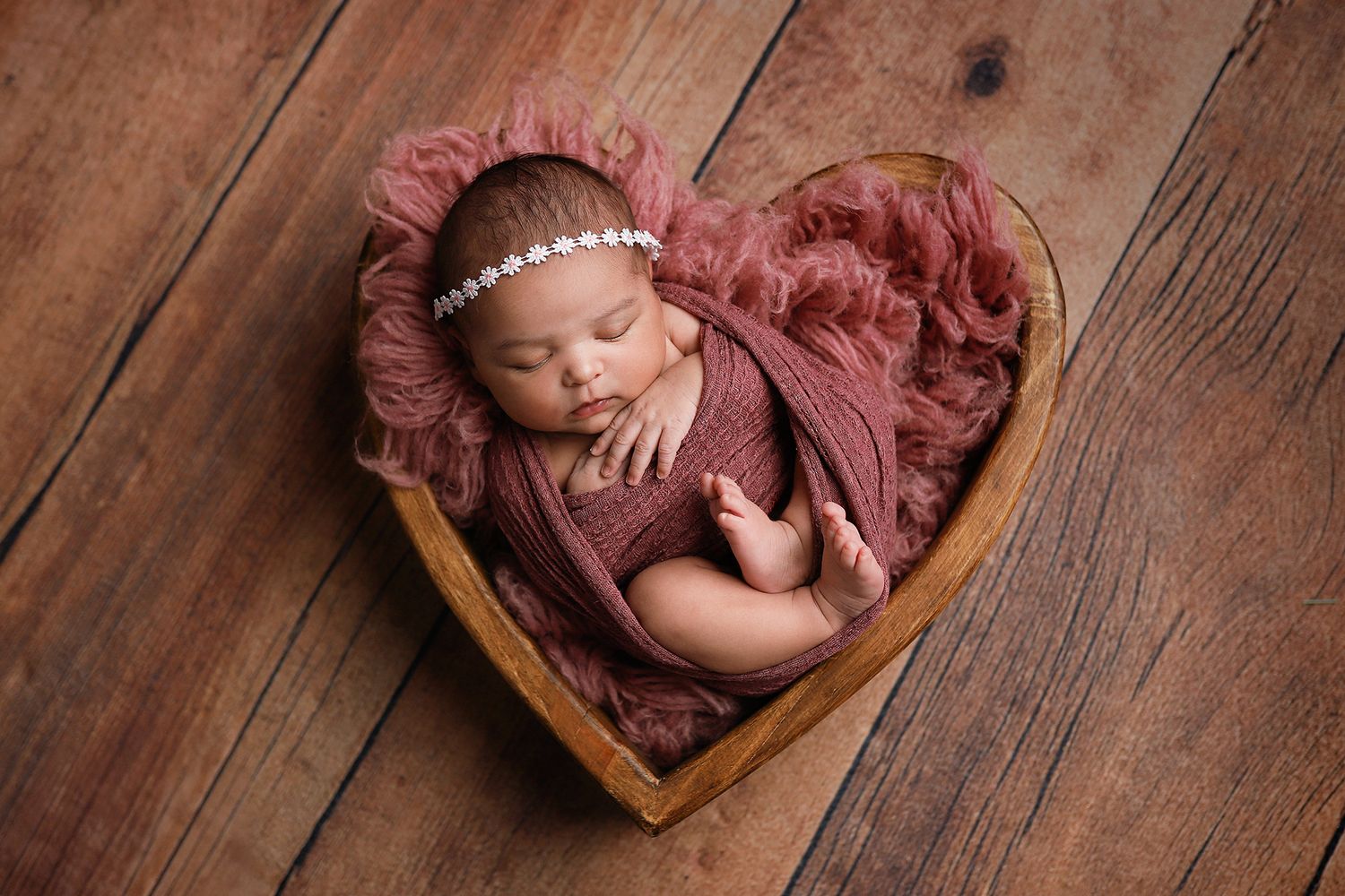 Newborn baby wrapped in dusty rose swaddle sleeping in wooden heart-shaped bowl with pearl headband.