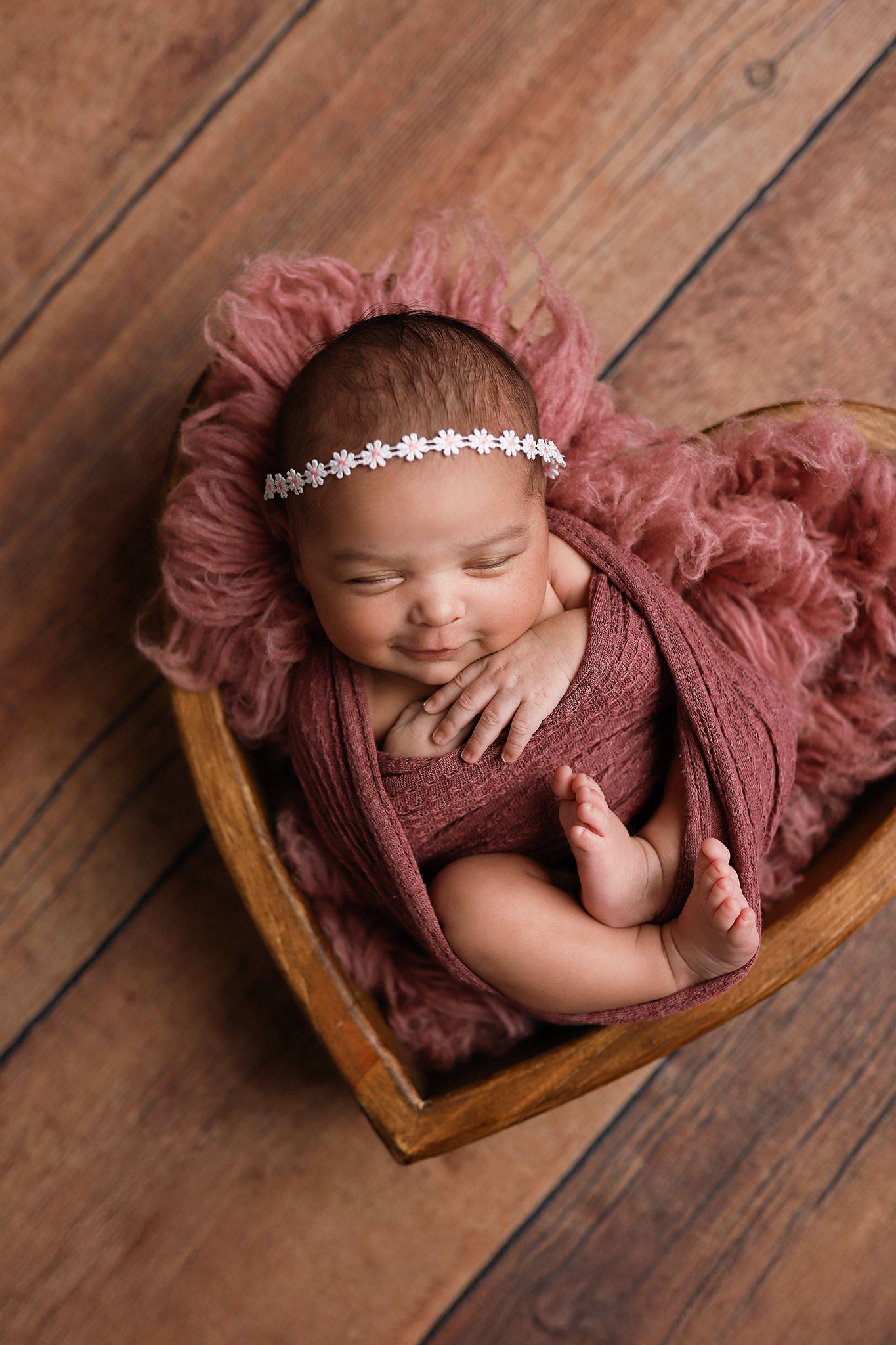 Smiling newborn baby wrapped in dusty rose swaddle with floral headband, sleeping in wooden heart-shaped bowl. Utah County newborn photos
