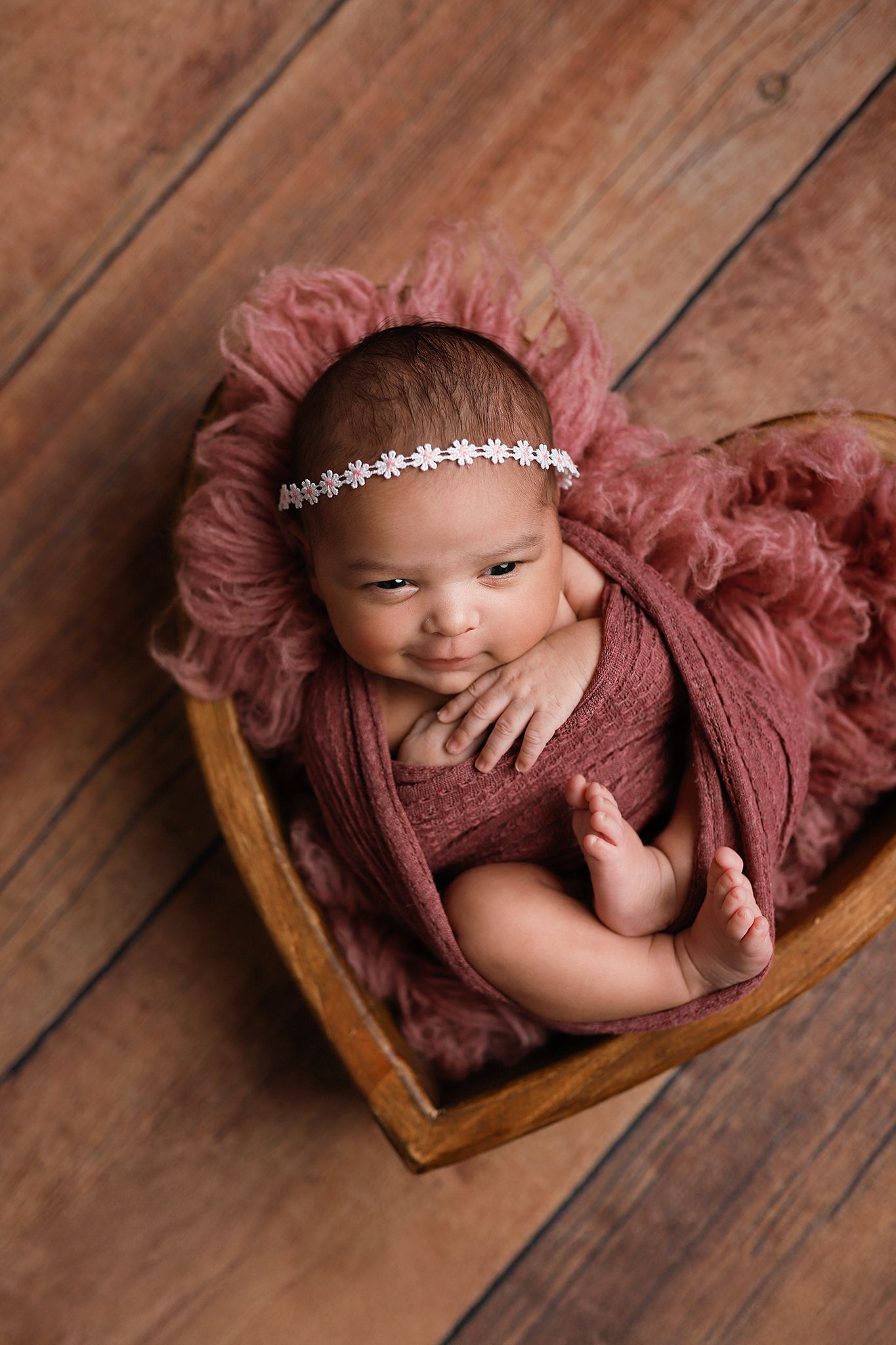 Newborn baby girl wrapped in dusty rose swaddle with floral headband, resting in heart-shaped wooden bowl on rustic wood floor.