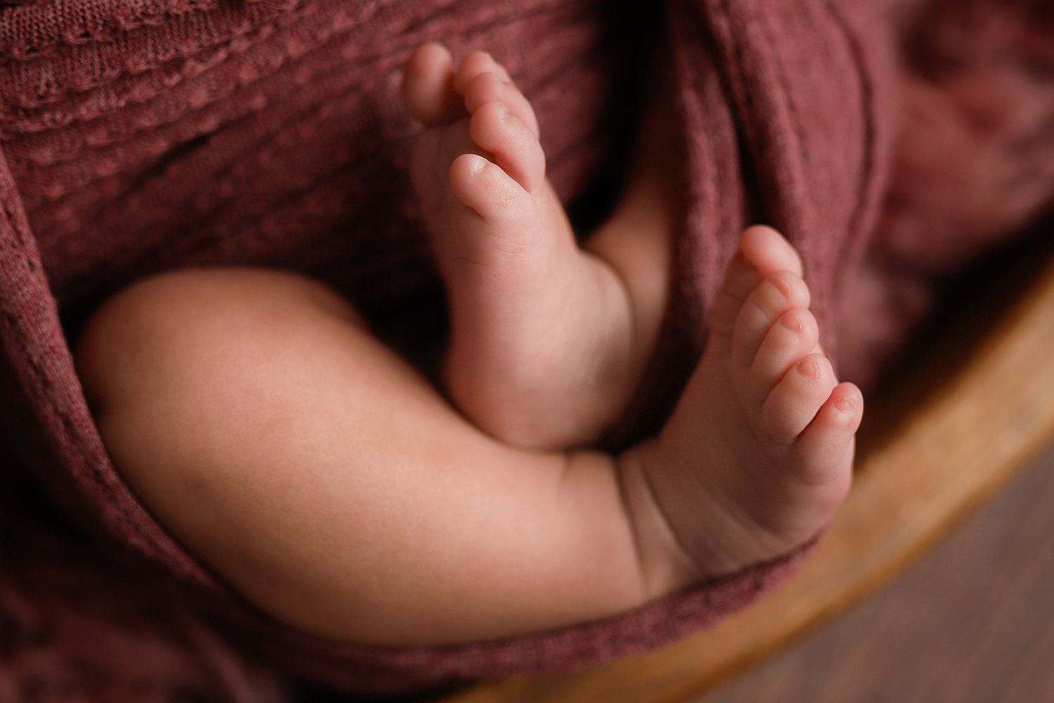 Close-up of newborn baby feet and legs wrapped in a soft mauve knit blanket inside a wooden bowl.