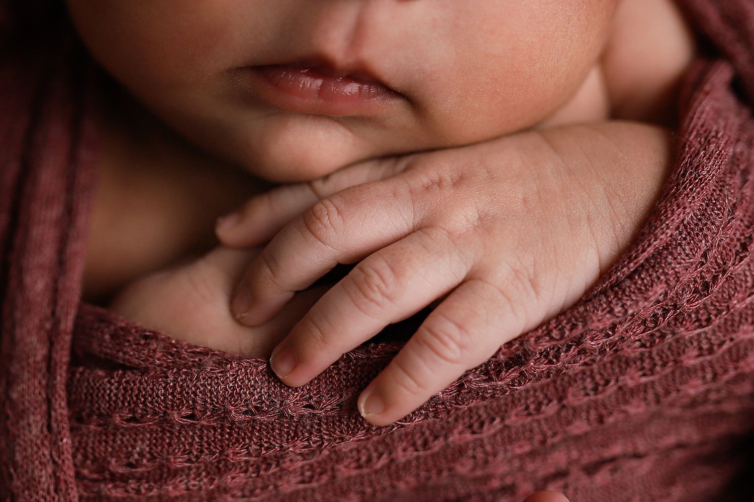 Close-up of a sleeping newborn baby wrapped in a soft pink knit blanket with tiny delicate fingers visible.