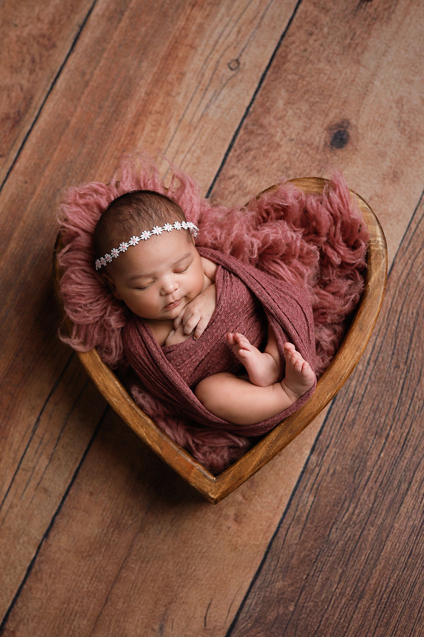 Sleeping newborn baby girl wrapped in dusty rose swaddle, resting in a wooden heart-shaped bowl with floral headband.