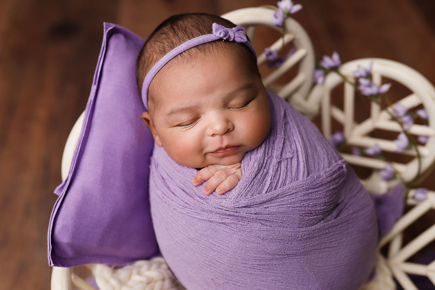 Newborn baby girl swaddled in purple wrap with bow headband, sleeping peacefully in white basket. Utah County newborn photographer