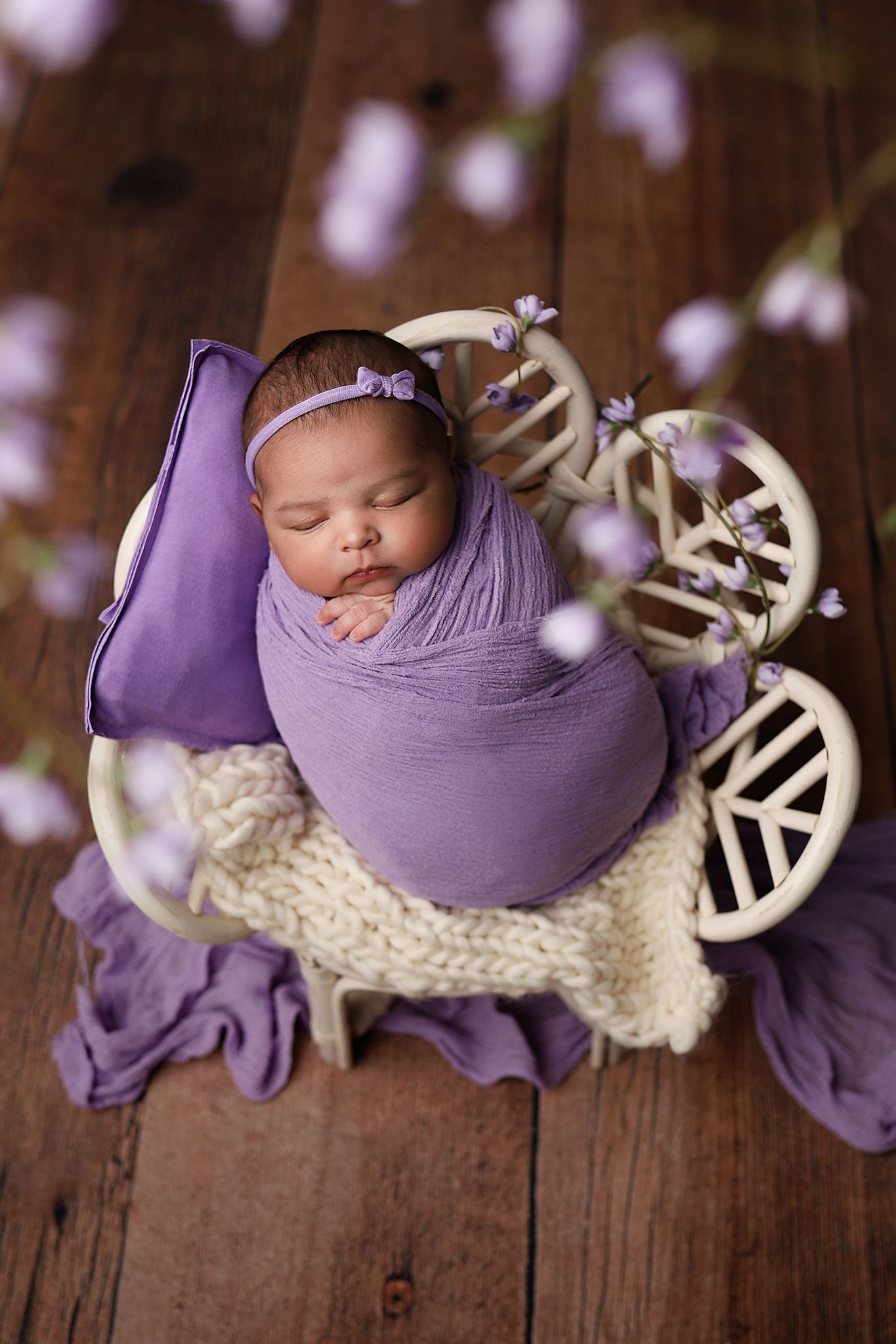 Sleeping newborn baby swaddled in purple wrap with floral headband, resting in a white bicycle prop on wooden floor. Utah newborn photos