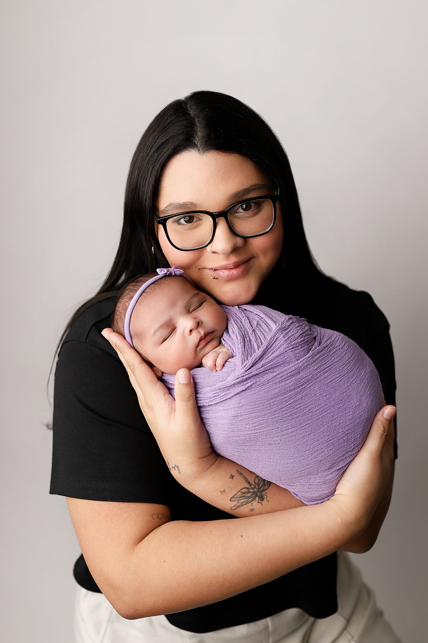 A young mother with black hair and glasses tenderly holds her swaddled newborn baby wrapped in purple cloth.
