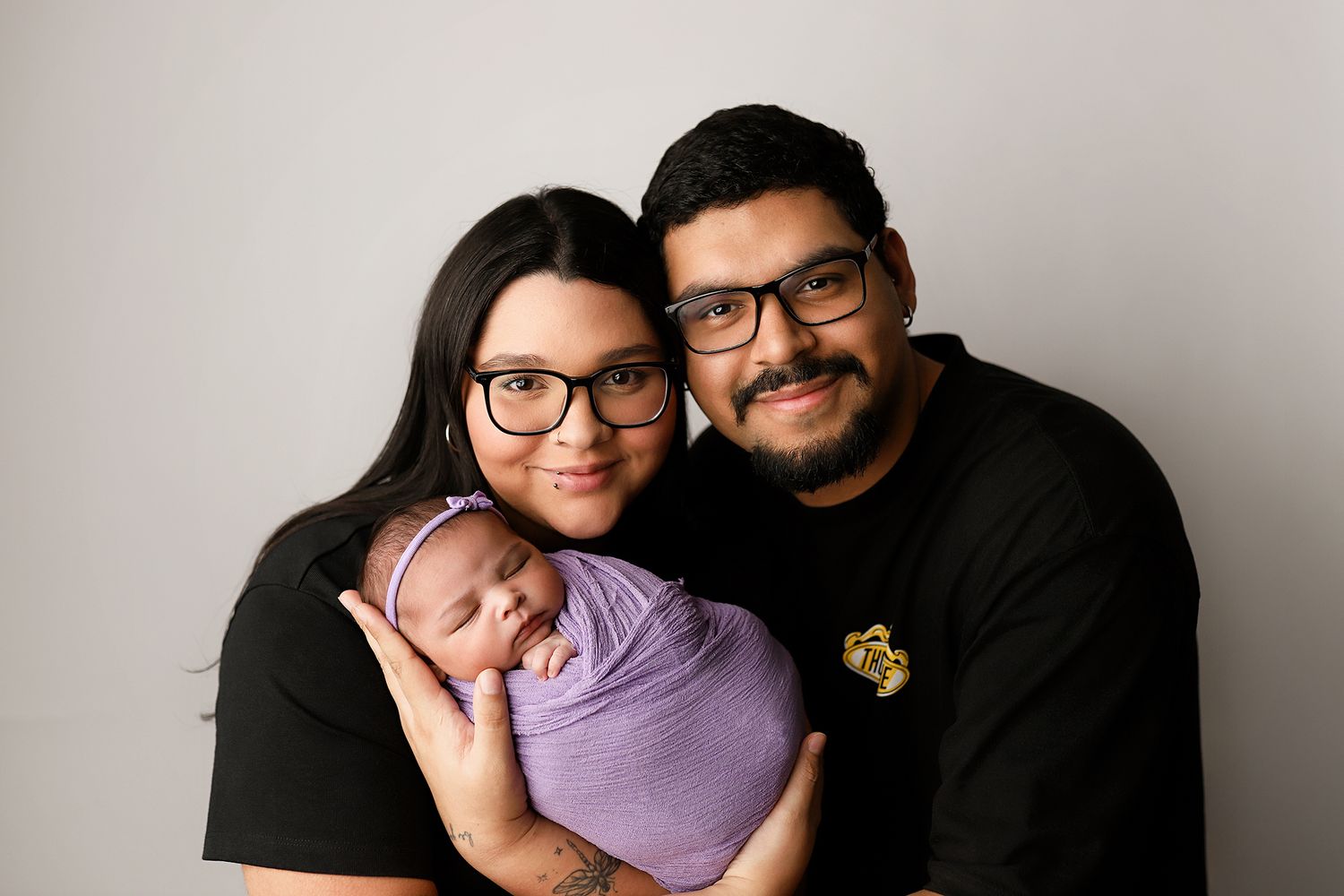 Parents in black shirts smiling and holding their newborn baby wrapped in purple for a family portrait.