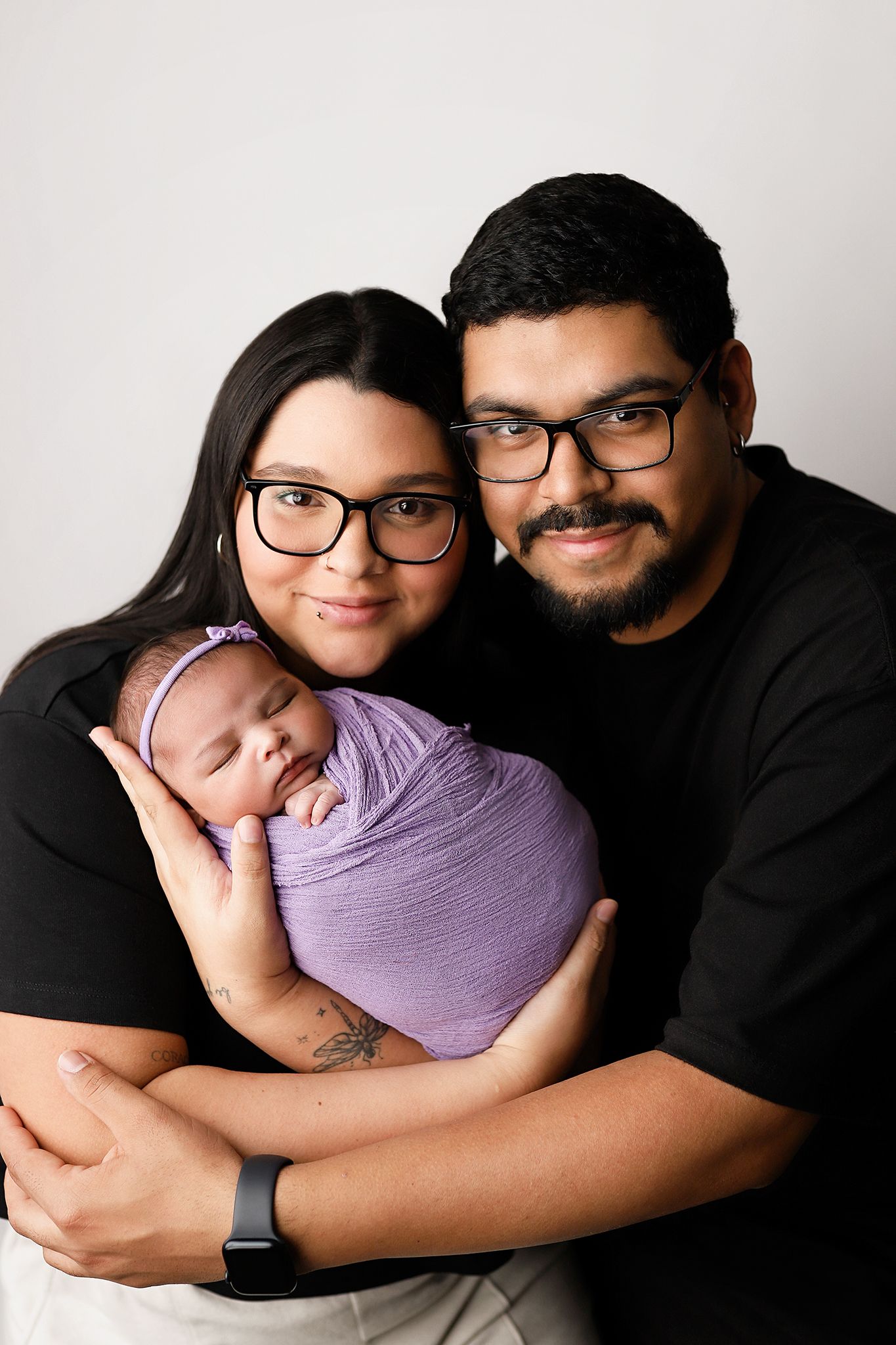 Young couple in black shirts and glasses holding their sleeping newborn baby wrapped in a purple swaddle.