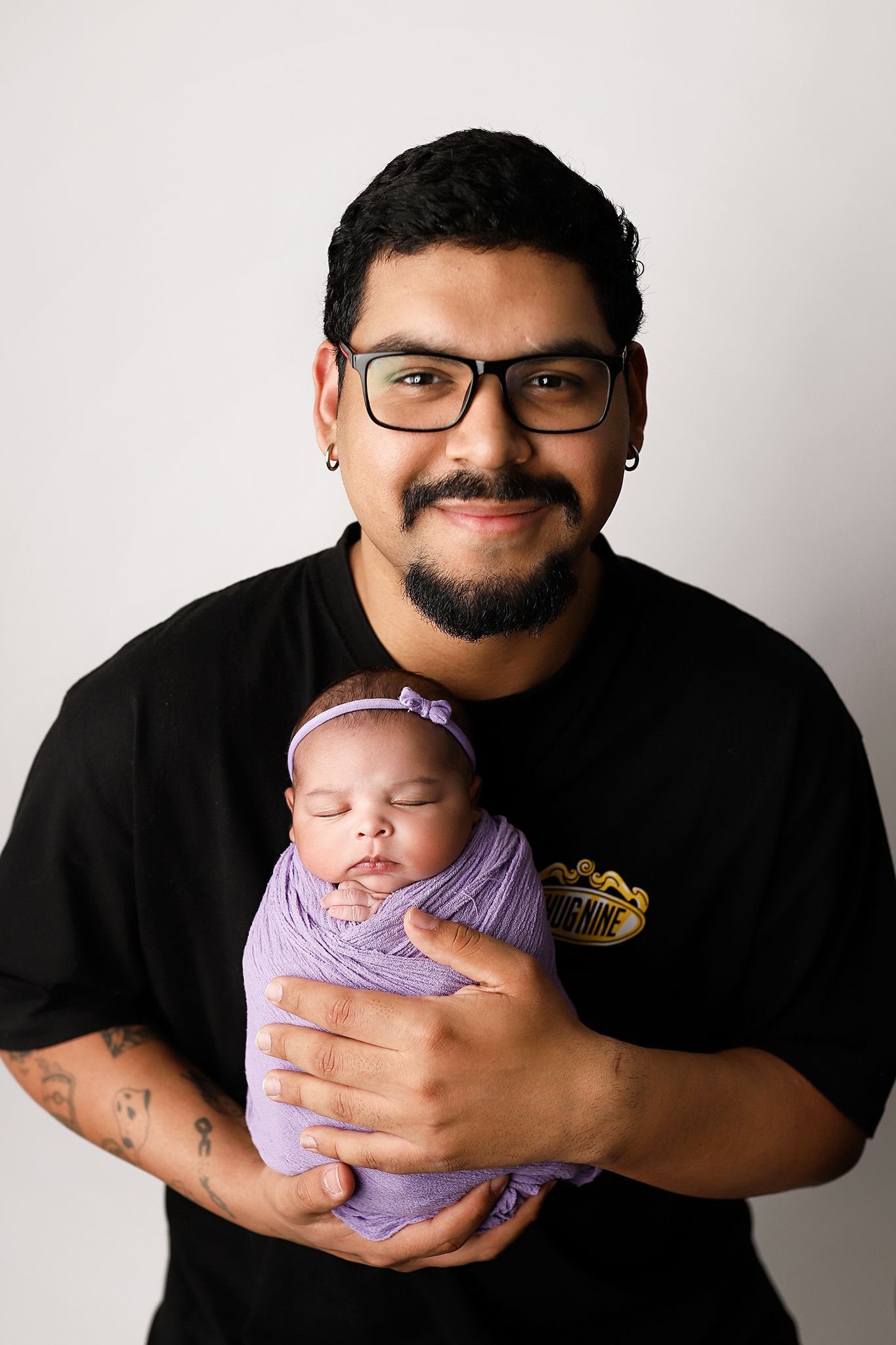 A smiling man with glasses and tattoos holds a sleeping newborn baby wrapped in a purple swaddle with a bow headband.
