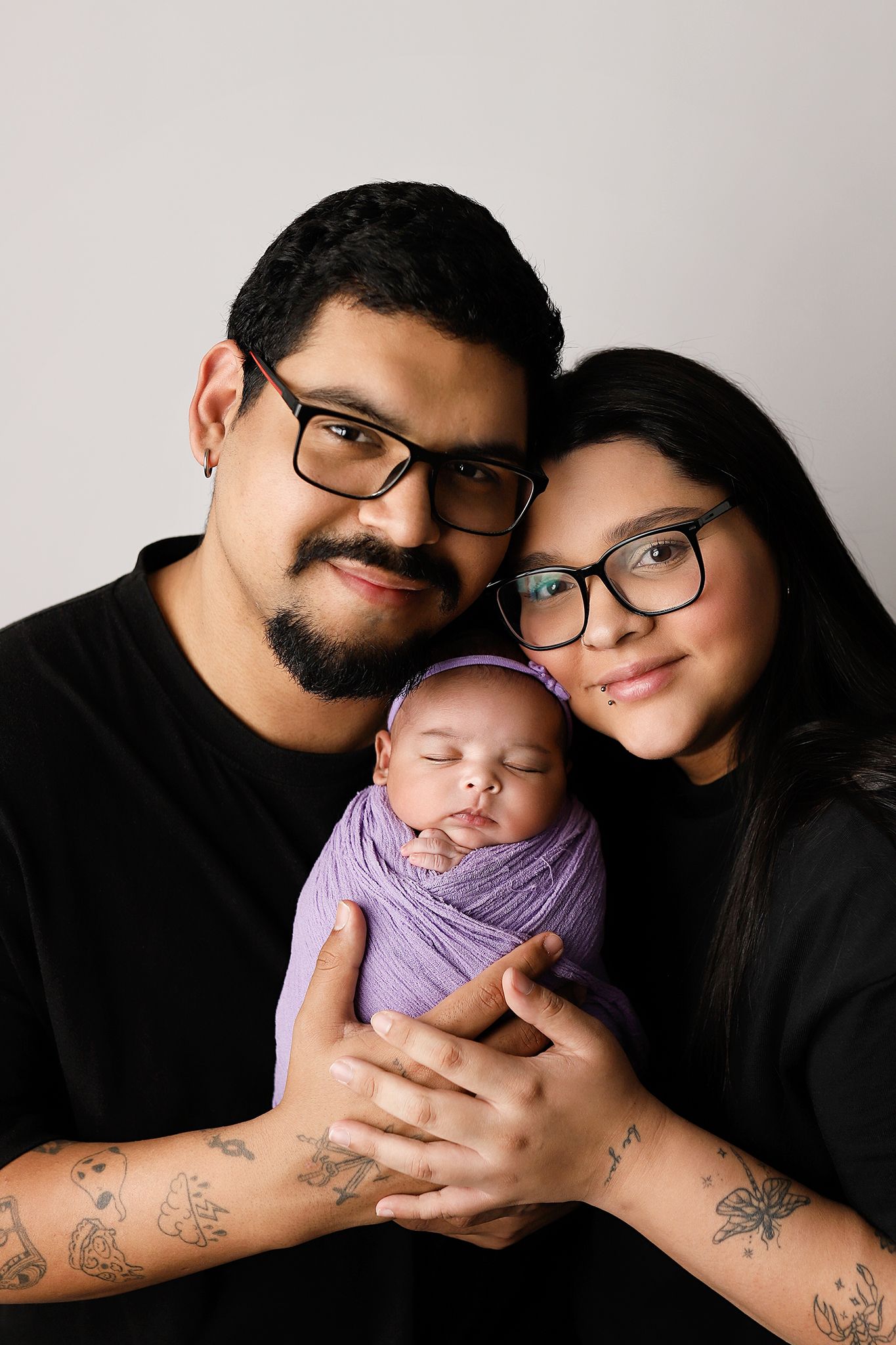 A young couple in black shirts and glasses tenderly hold their swaddled newborn in a purple wrap for a studio portrait.