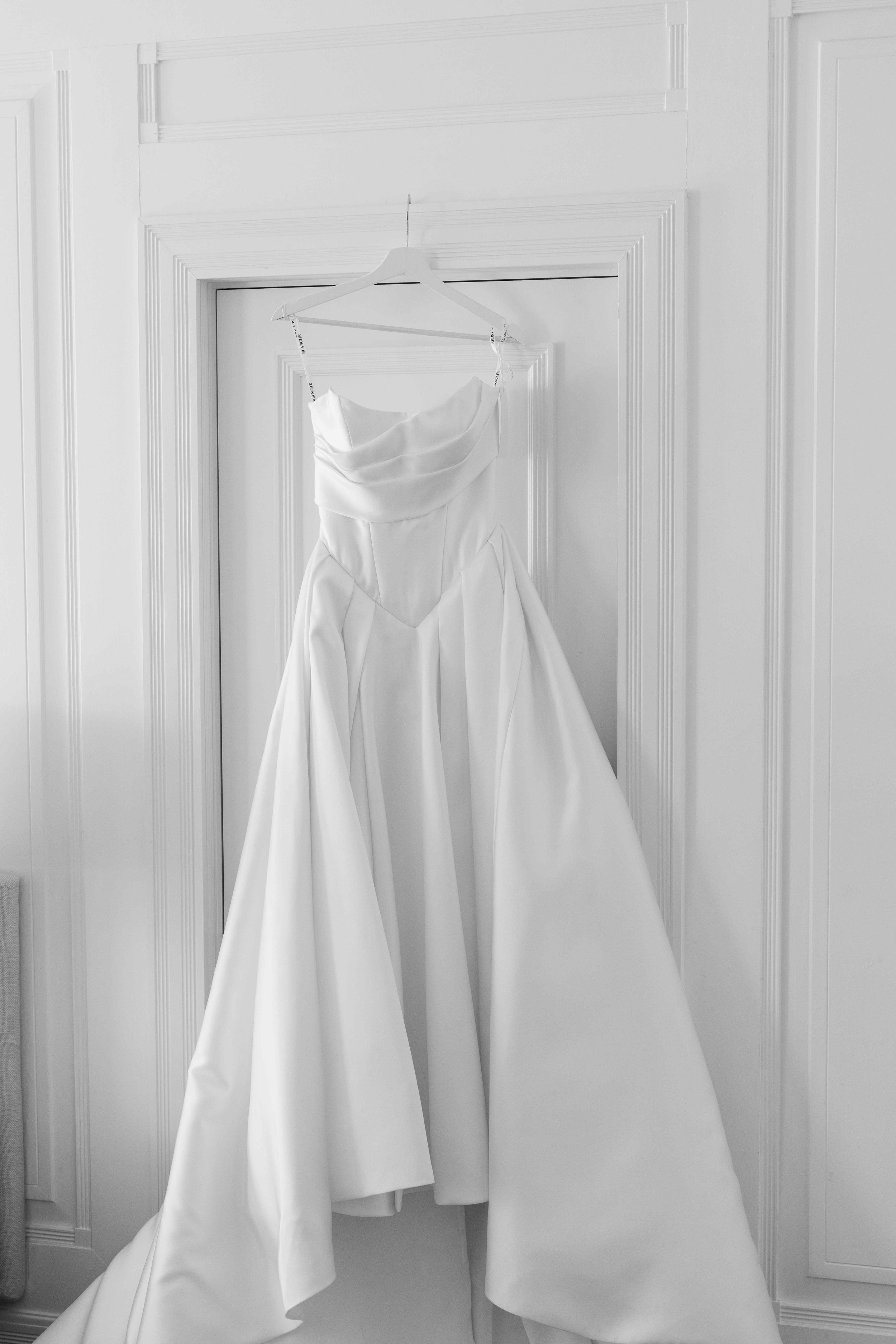 Elegant white strapless wedding dress hanging on a hanger against a white paneled wall in black and white photo.