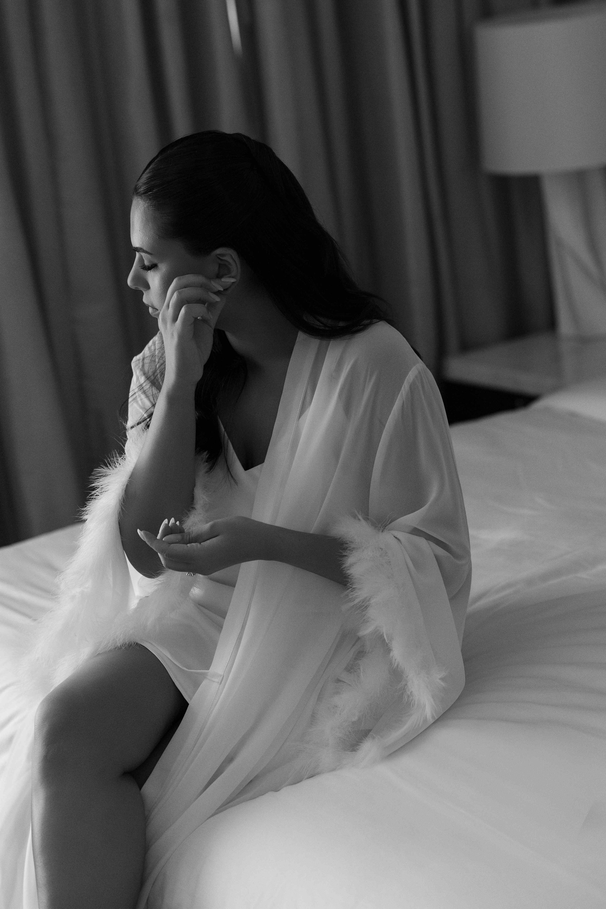 Emotional bride in white feathered robe sitting on bed holding jewelry, candid black and white wedding morning photo.
