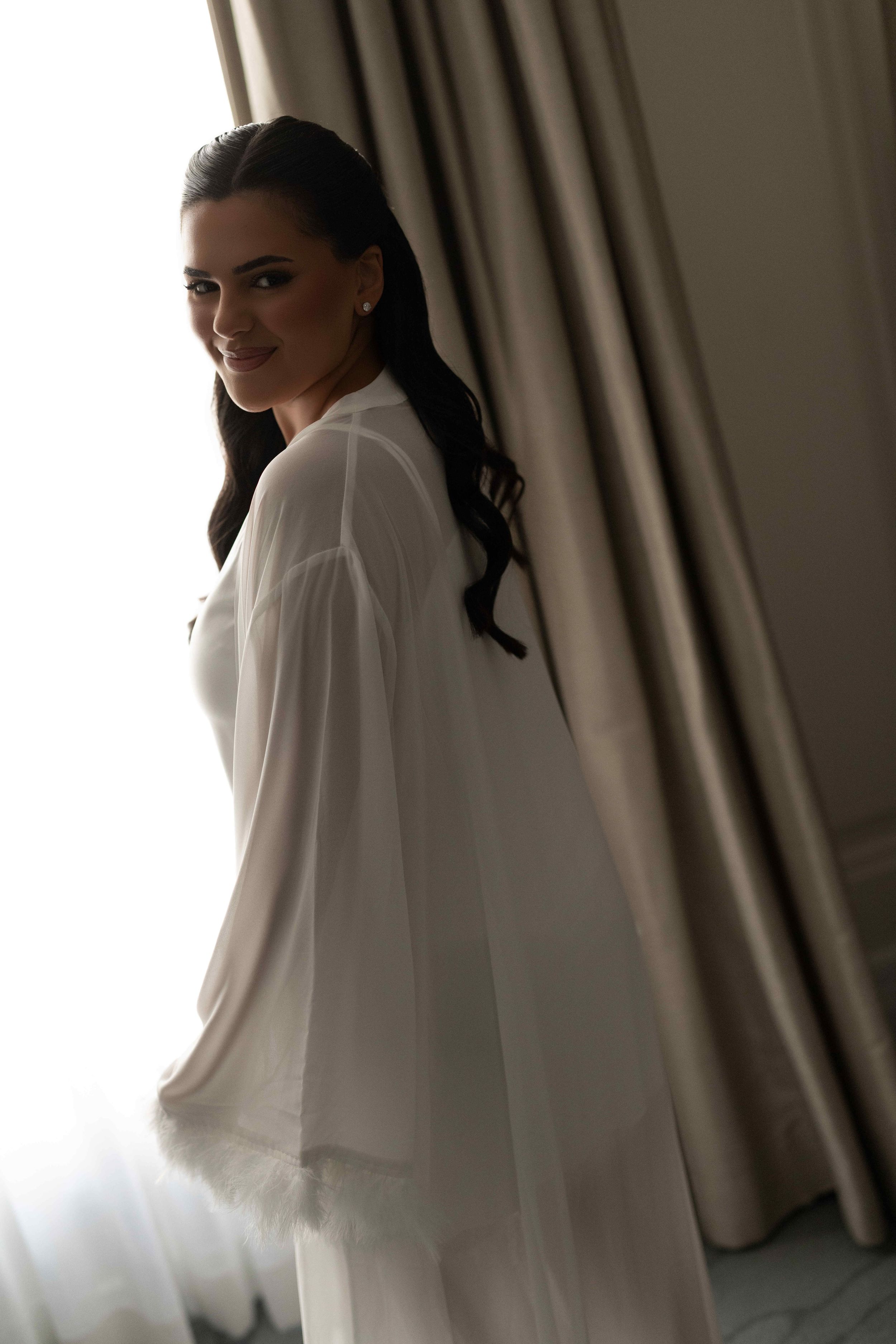 A smiling brunette woman in a white flowing robe stands elegantly near a curtained window in soft natural light.