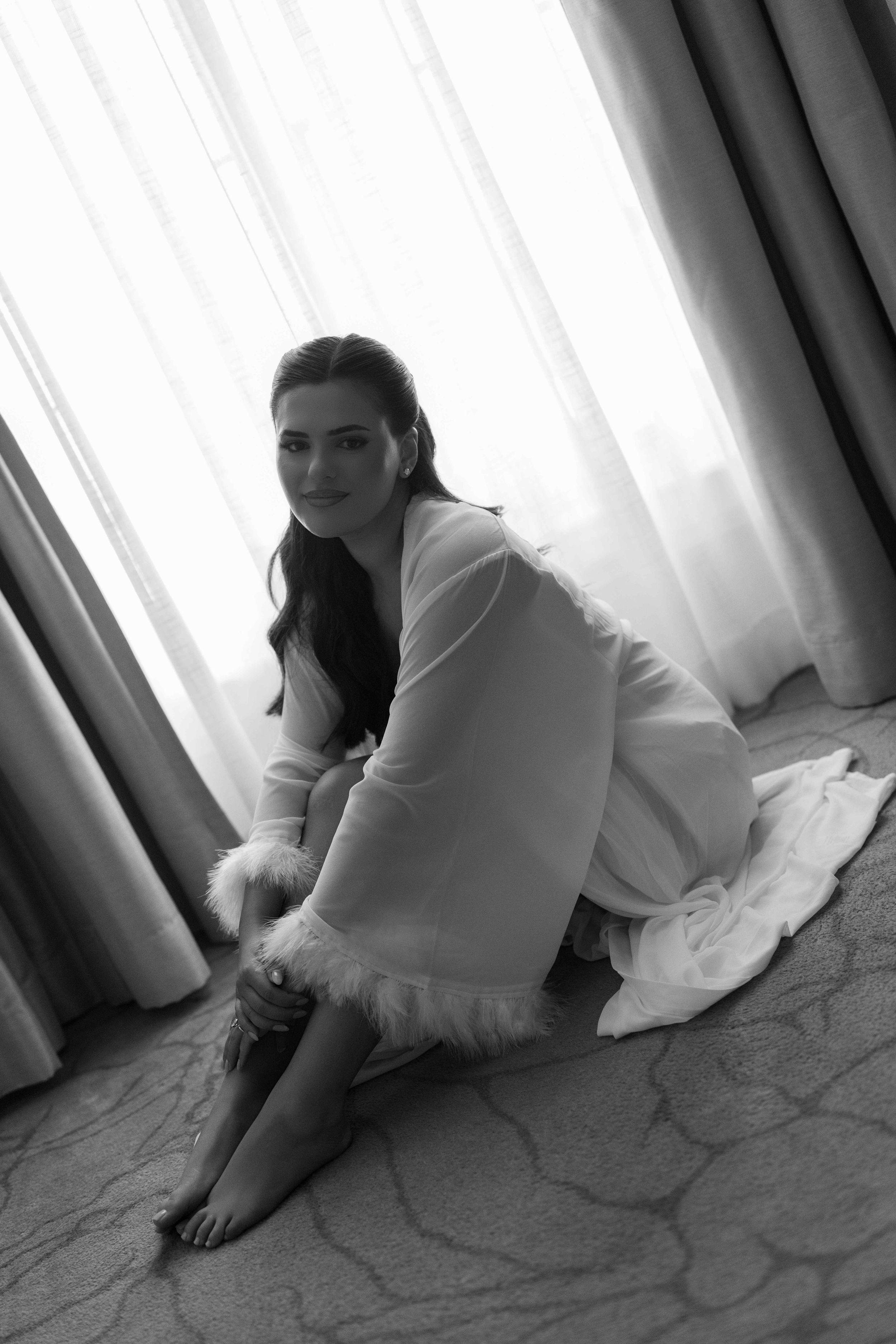 Black and white photo of a smiling woman in a white robe with fur trim sitting on a carpeted floor near curtained windows.