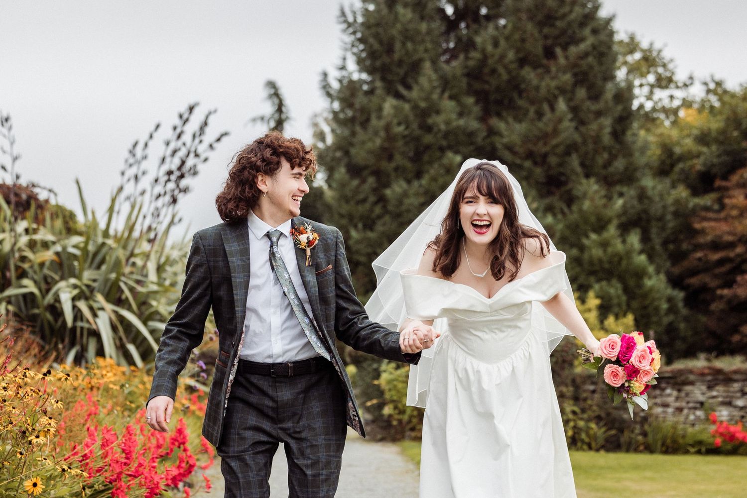 Joyful bride and groom holding hands running through a garden, bride in white gown with veil holding pink bouquet. Cragwood Hotel.