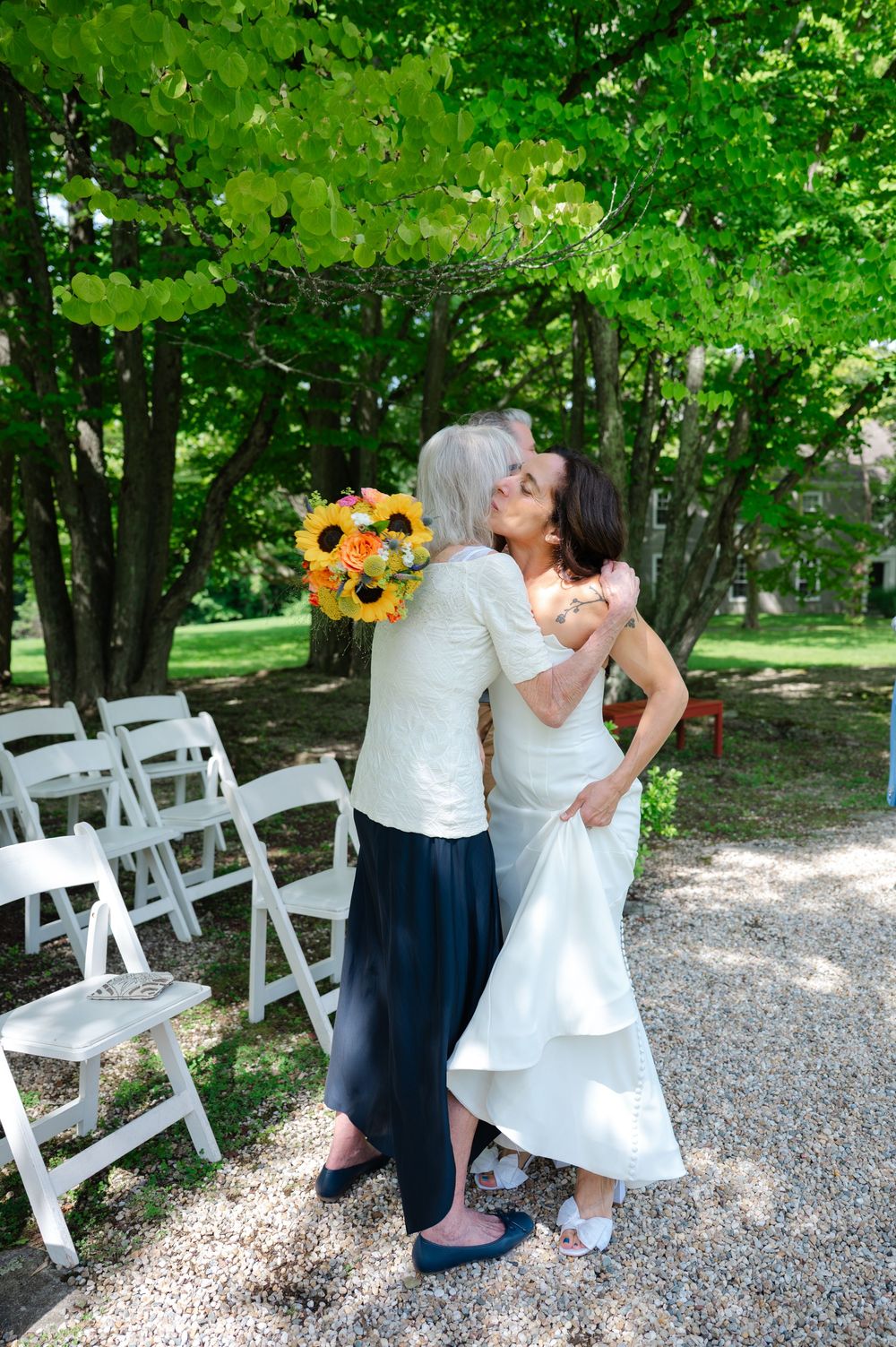 Bride in white gown hugging older woman holding sunflower bouquet outdoors at wedding ceremony with white chairs.