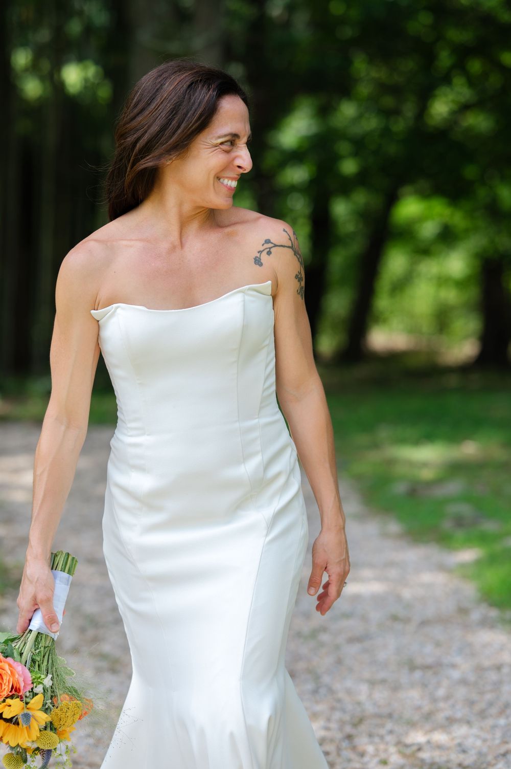 Smiling bride in strapless white mermaid gown walks outdoors holding colorful bouquet, shoulder tattoo visible.