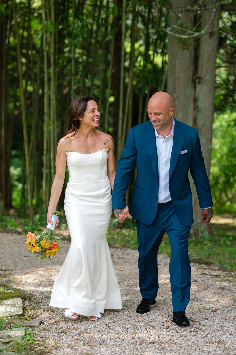 Bride in white gown and groom in blue suit walk hand-in-hand on a gravel path surrounded by lush green trees.