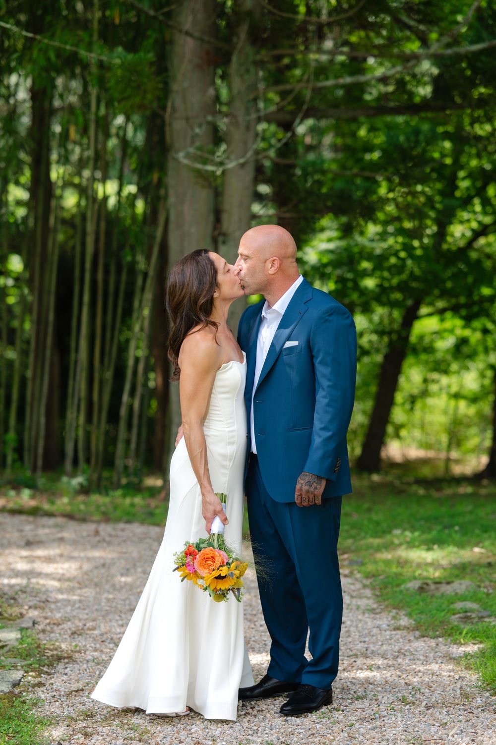 Bride and groom sharing a kiss on a gravel path surrounded by lush green trees, bride holding vibrant orange bouquet.