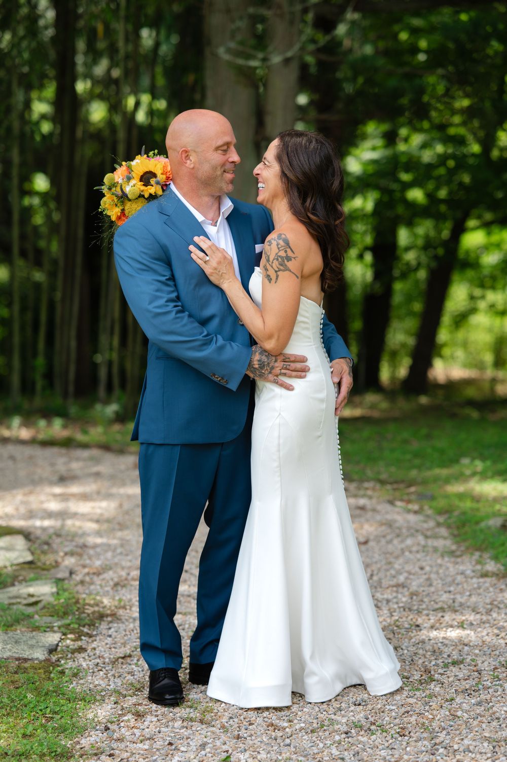 Bride and groom embrace on a gravel path surrounded by lush greenery, bride holding sunflower bouquet in white gown.