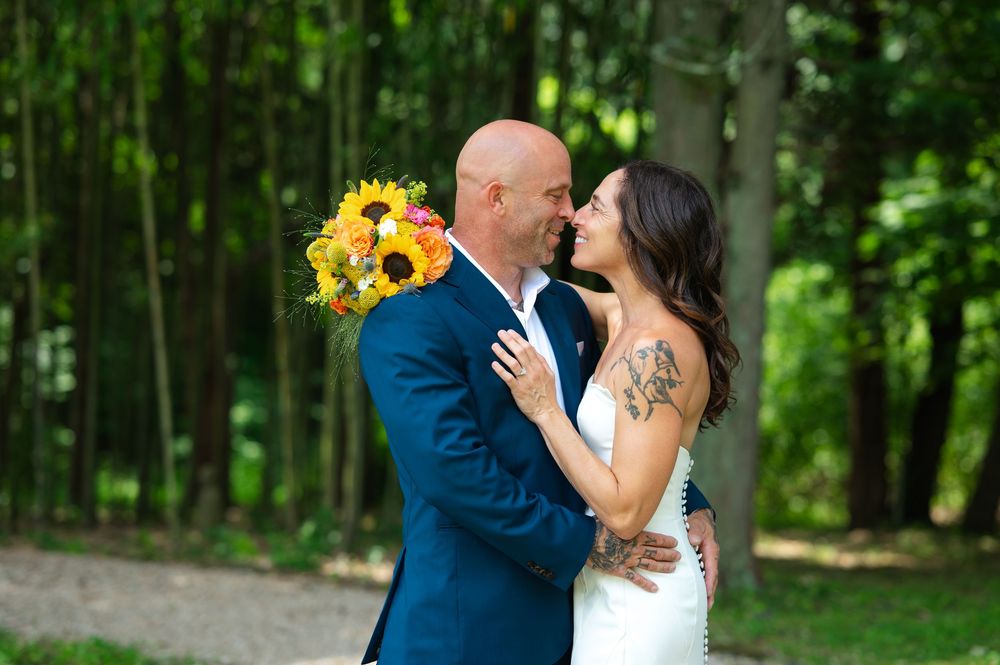 Bride and groom share a romantic moment outdoors, with sunflower bouquet, lush green trees in background.
